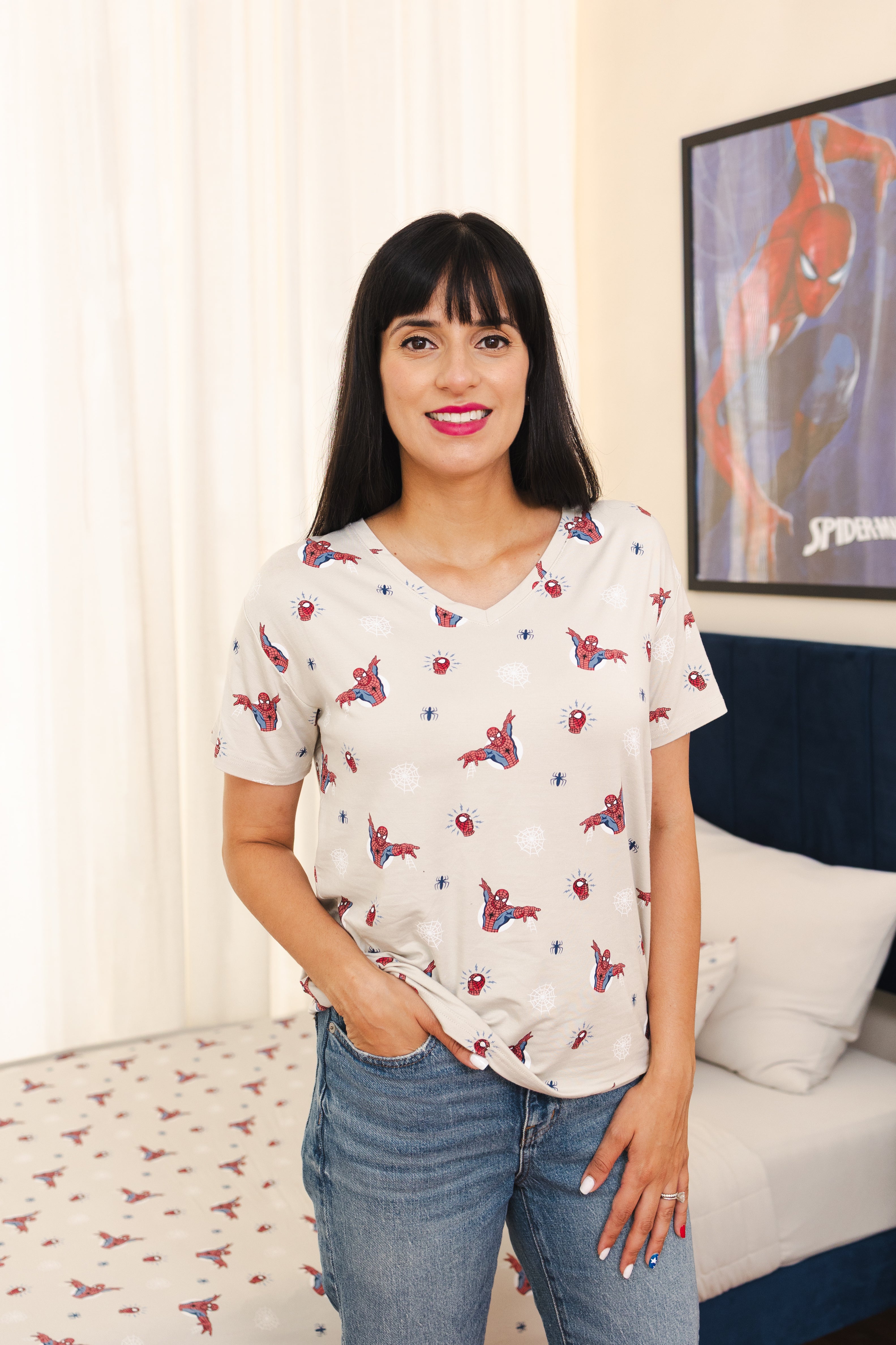 A woman wearing a short-sleeve white t-shirt with a red and blue Spider-Man print, paired with blue jeans, standing in a bedroom setting.