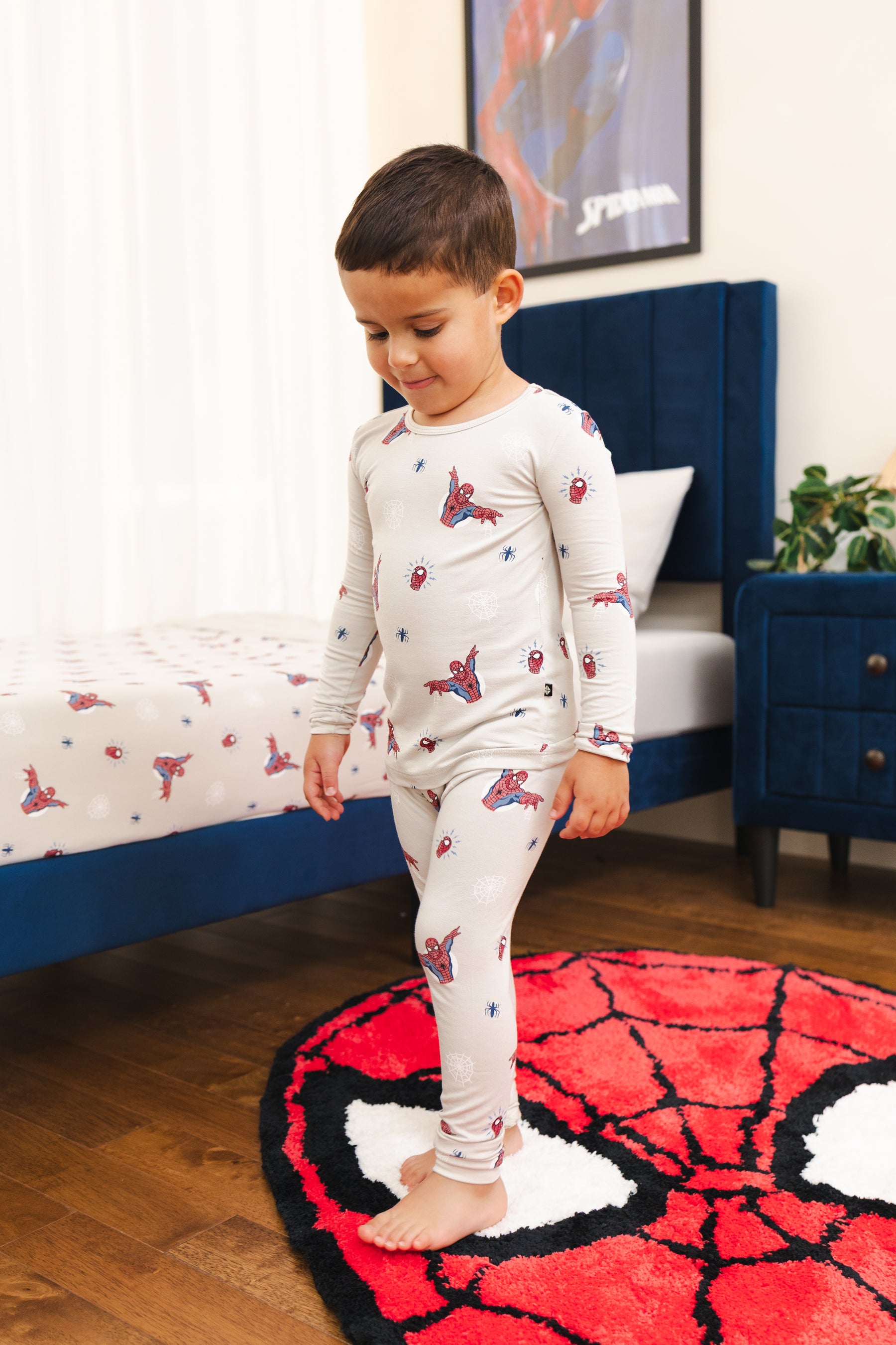A young boy wearing a light beige long-sleeve pajama set with a red and blue Spider-Man print, standing barefoot on a red and black Spider-Man themed rug in a bedroom.