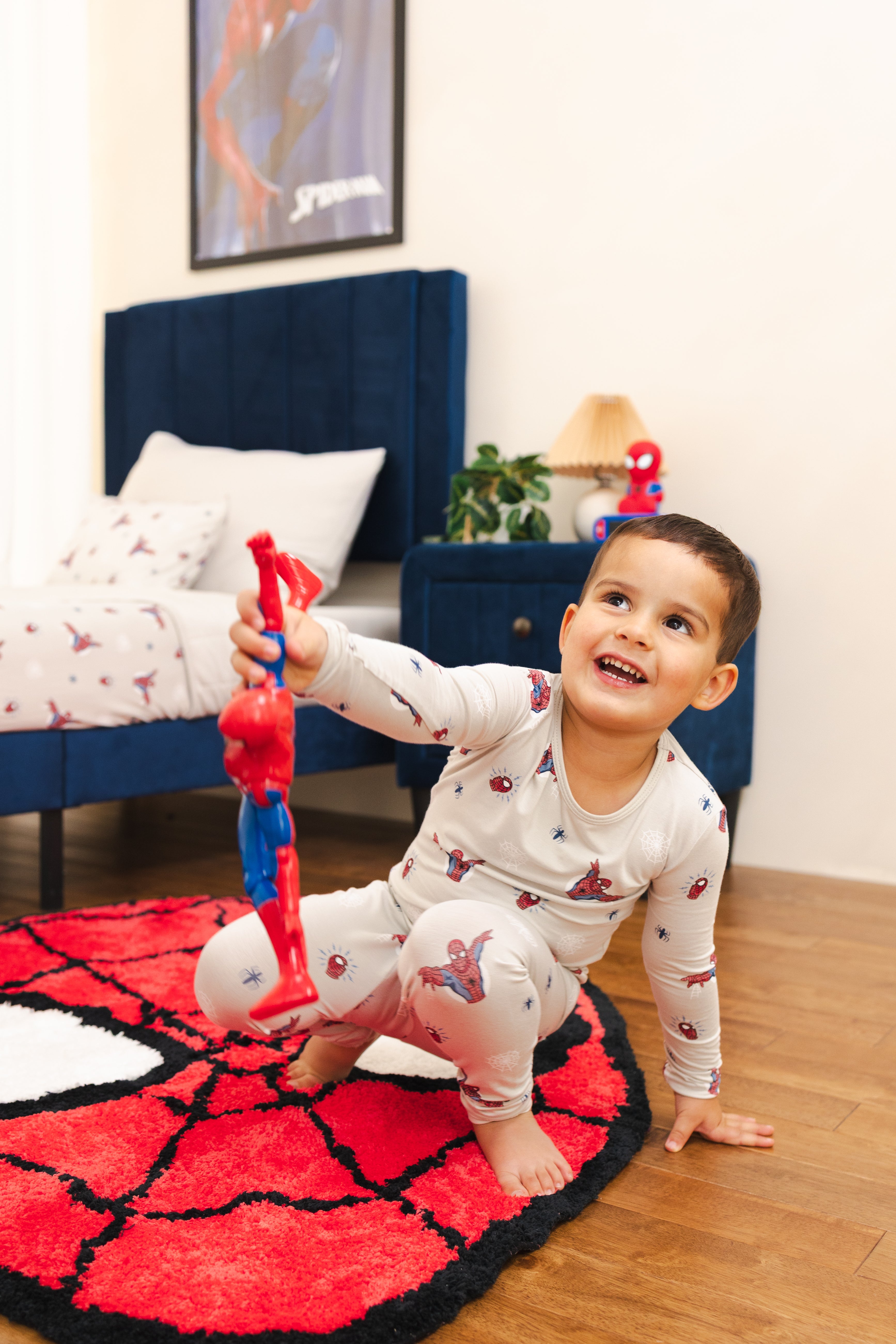 A young boy wearing a Spider-Man themed pajama set crawls on a red and black Spider-Man themed rug in a bedroom, holding a Spider-Man toy.