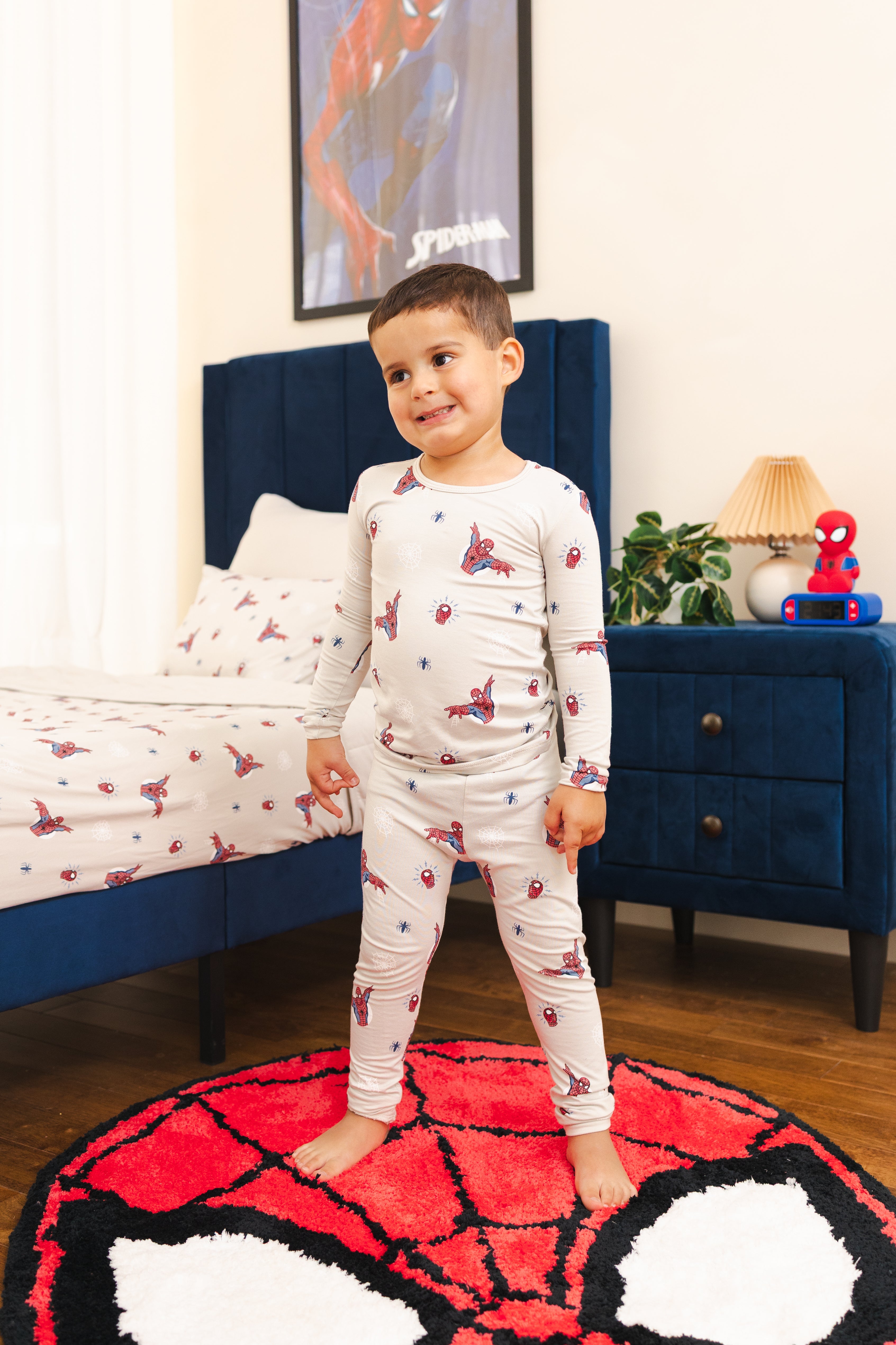 A young boy stands barefoot on a round red, white, and black Spider-Man-themed rug in a bedroom, wearing a light gray long-sleeve pajama set printed with red and blue Spider-Man web motifs. The room features a dark blue bed frame, matching headboard, and a blue nightstand with a Spider-Man figure and lamp.