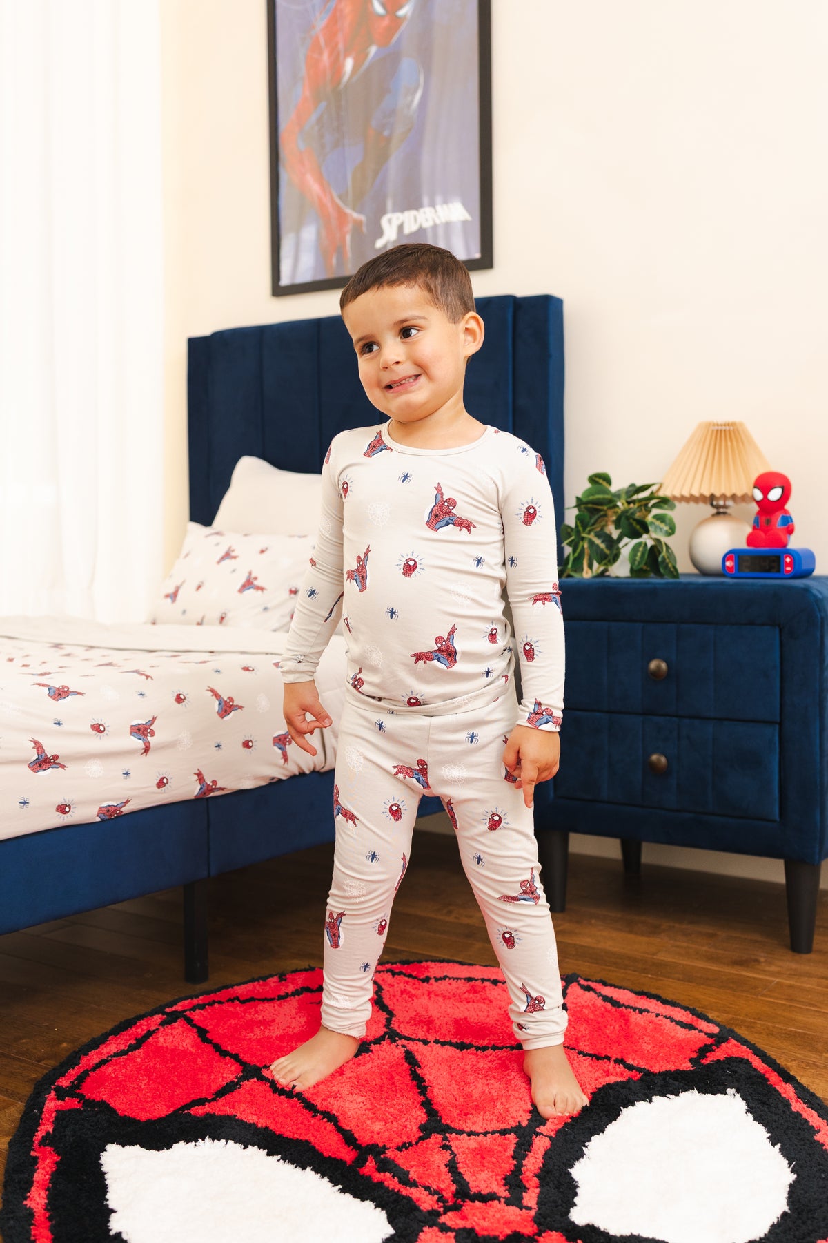 A young boy stands barefoot on a round red, white, and black Spider-Man-themed rug in a bedroom, wearing a light gray long-sleeve pajama set printed with red and blue Spider-Man web motifs. The room features a dark blue bed frame, matching headboard, and a blue nightstand with a Spider-Man figure and lamp.