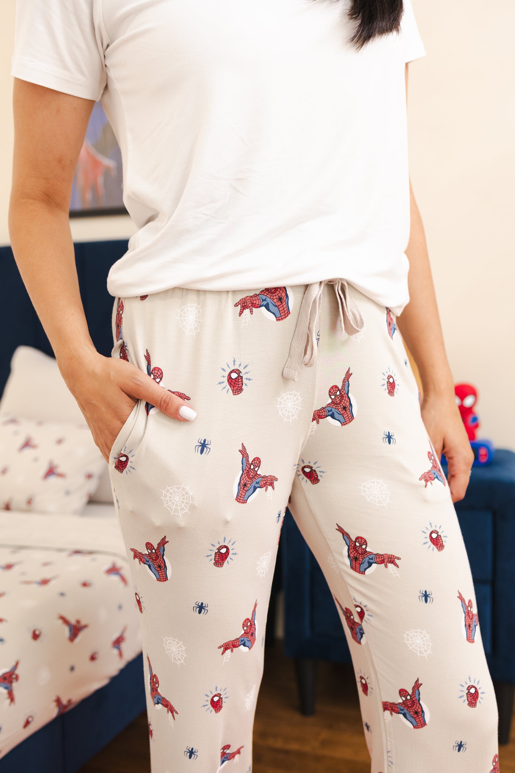 A person wearing a white t-shirt and cream-colored pajama pants with a Spider-Man print, standing in a bedroom setting.