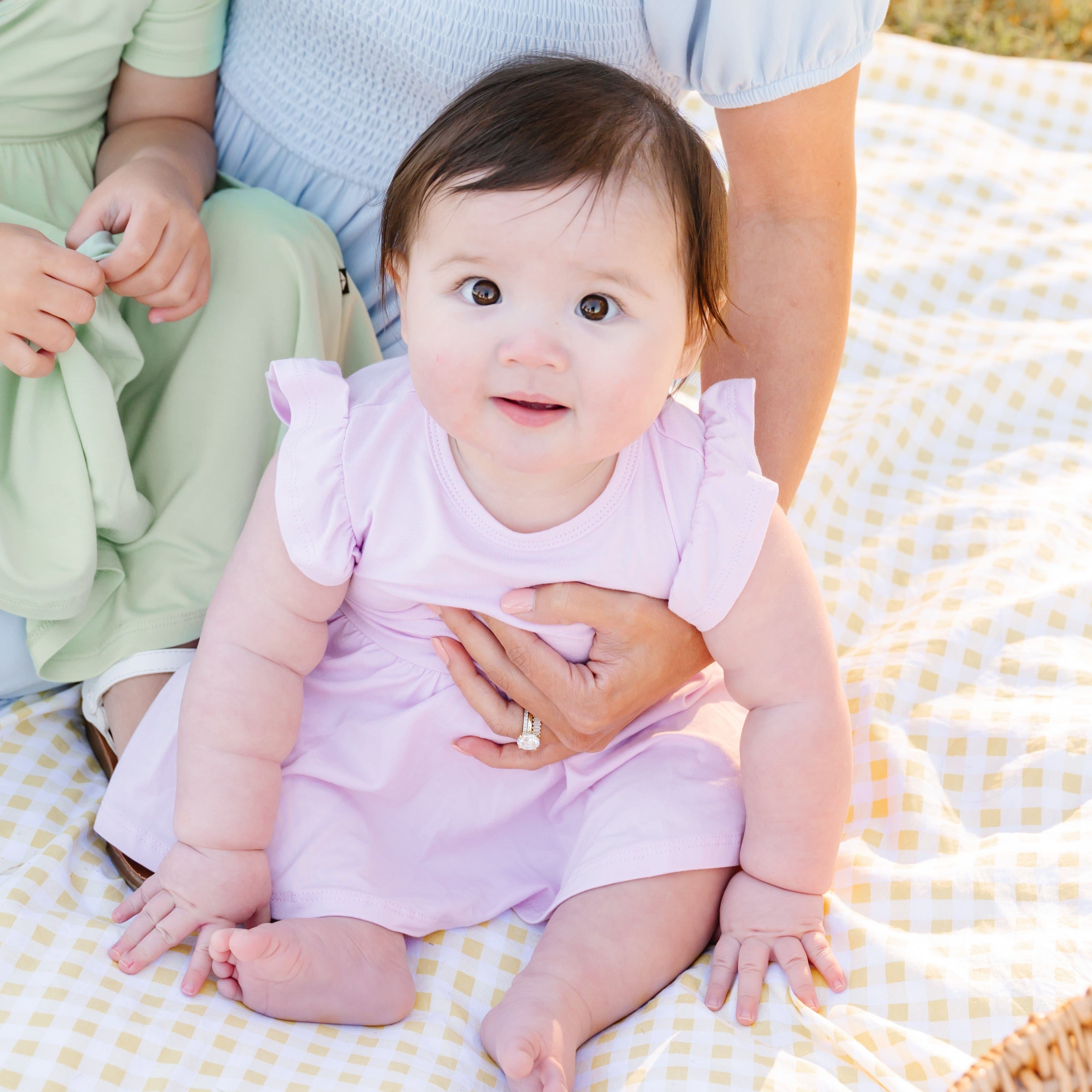 Baby in a pink twirl bodysuit dress sitting on a yellow checkered blanket with adults partially visible.