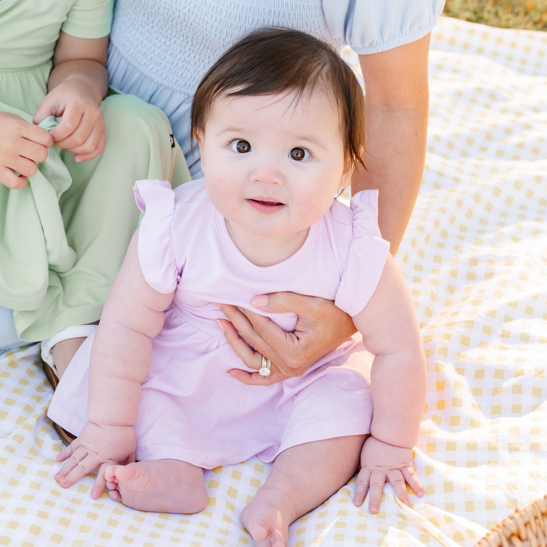 Baby in a pink twirl bodysuit dress sitting on a yellow checkered blanket with adults partially visible.