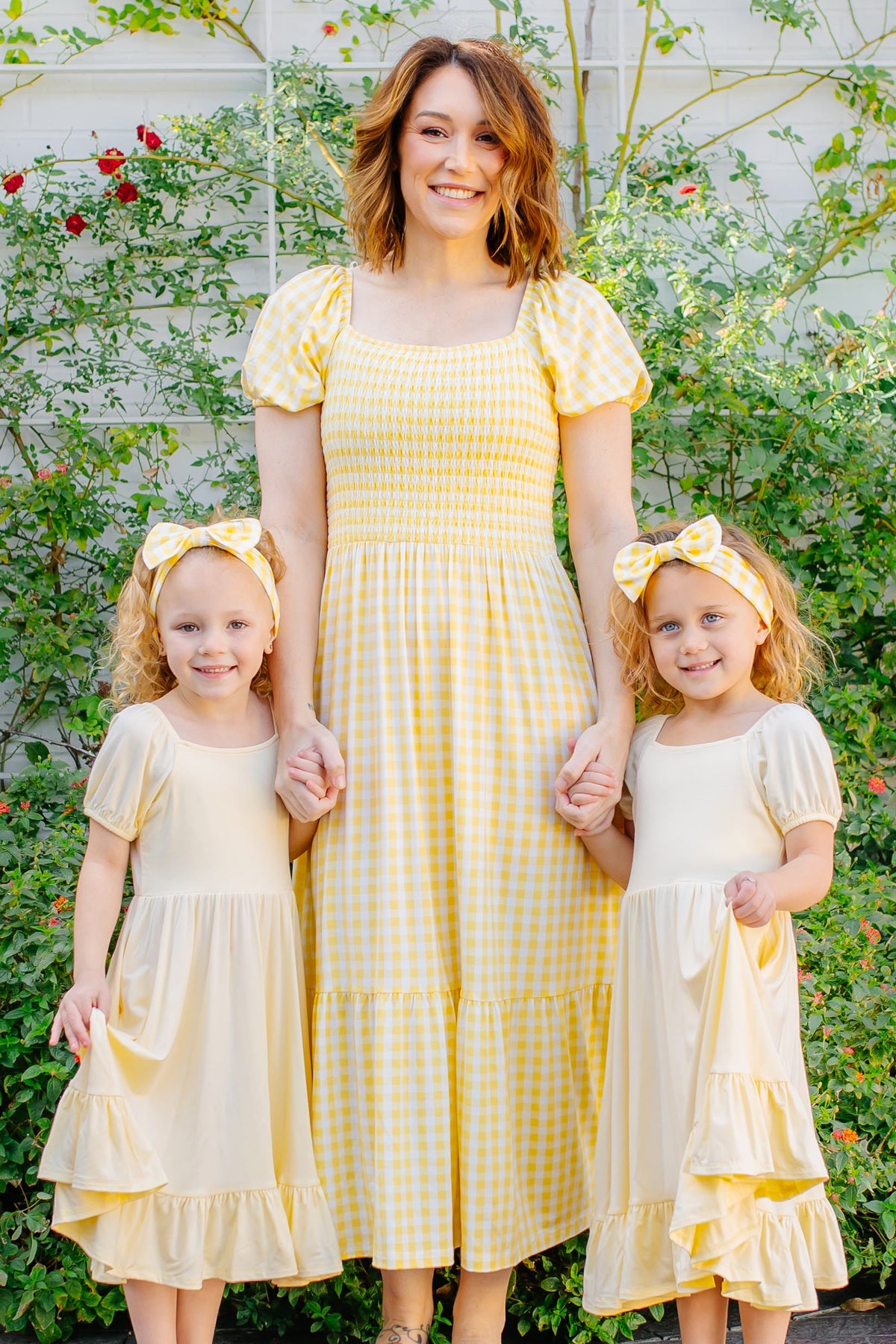 Woman and two young girls in matching yellow dresses standing outdoors with greenery in the background