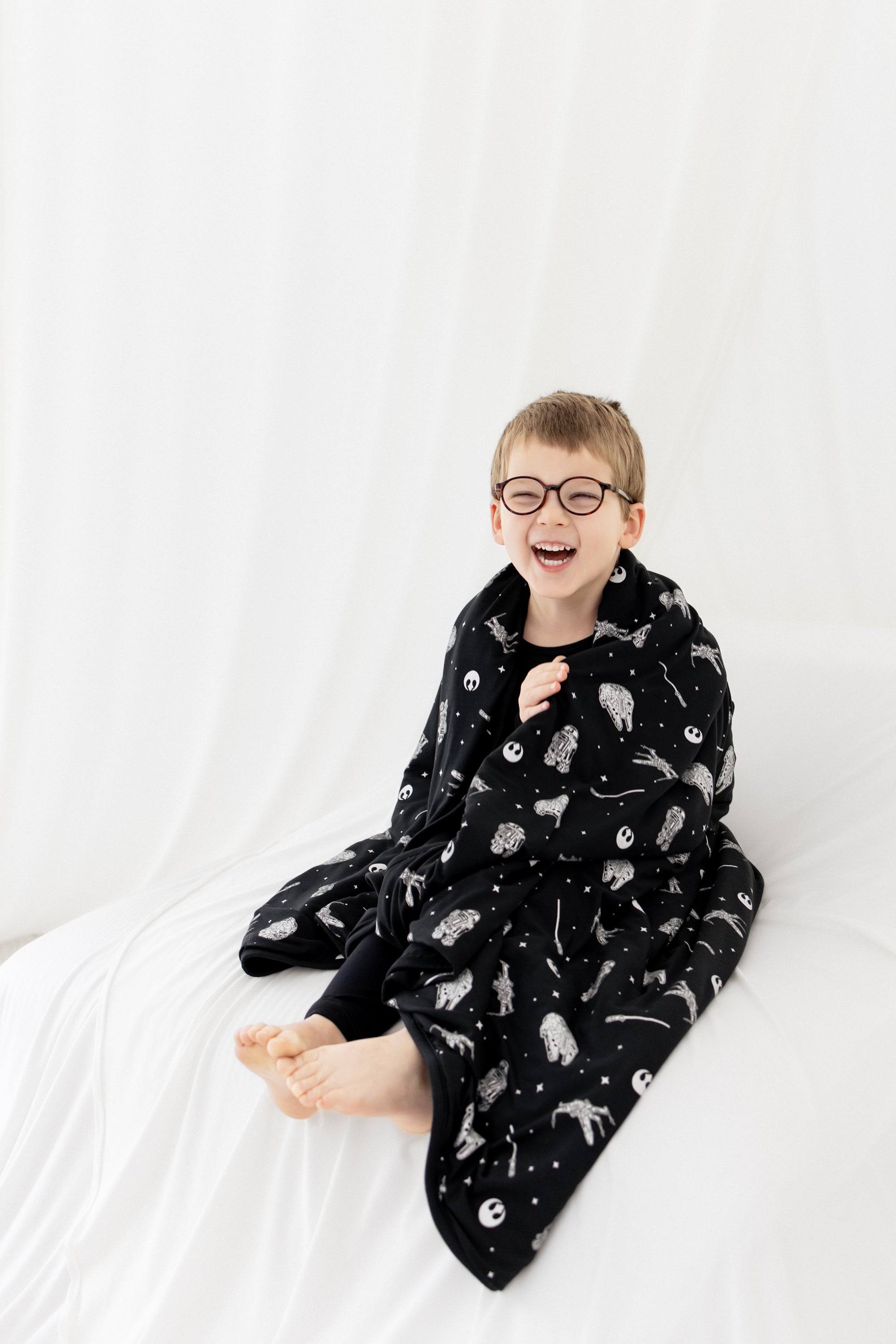 Young smiling boy sitting on a white blanket with the Toddler Blanket in STAR WARS™ 1.0 around his shoulders