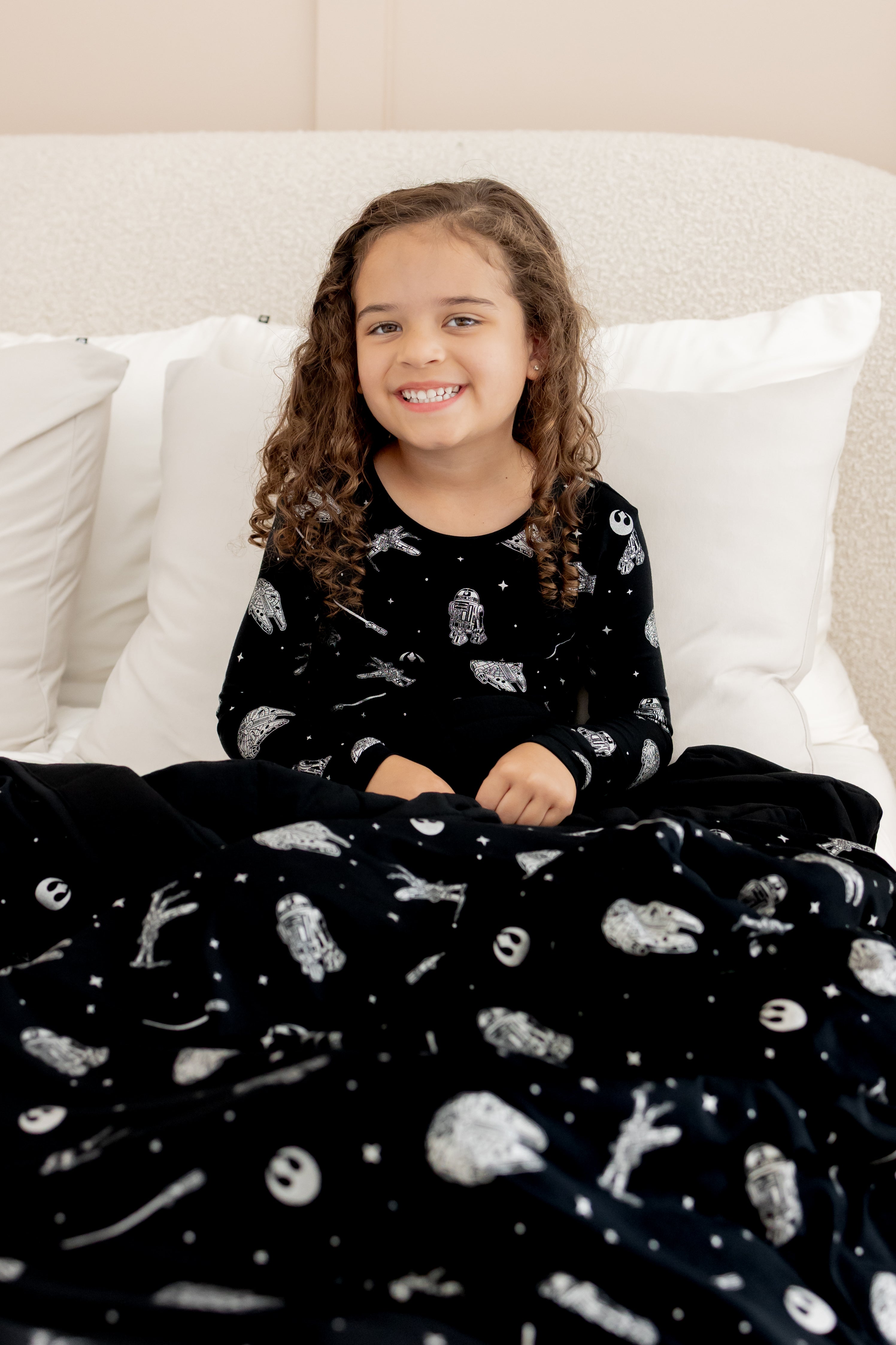 Girl sitting on bed covered under Youth Blanket in STAR WARS™ 2.5 print wearing similar print long sleeve pajama set