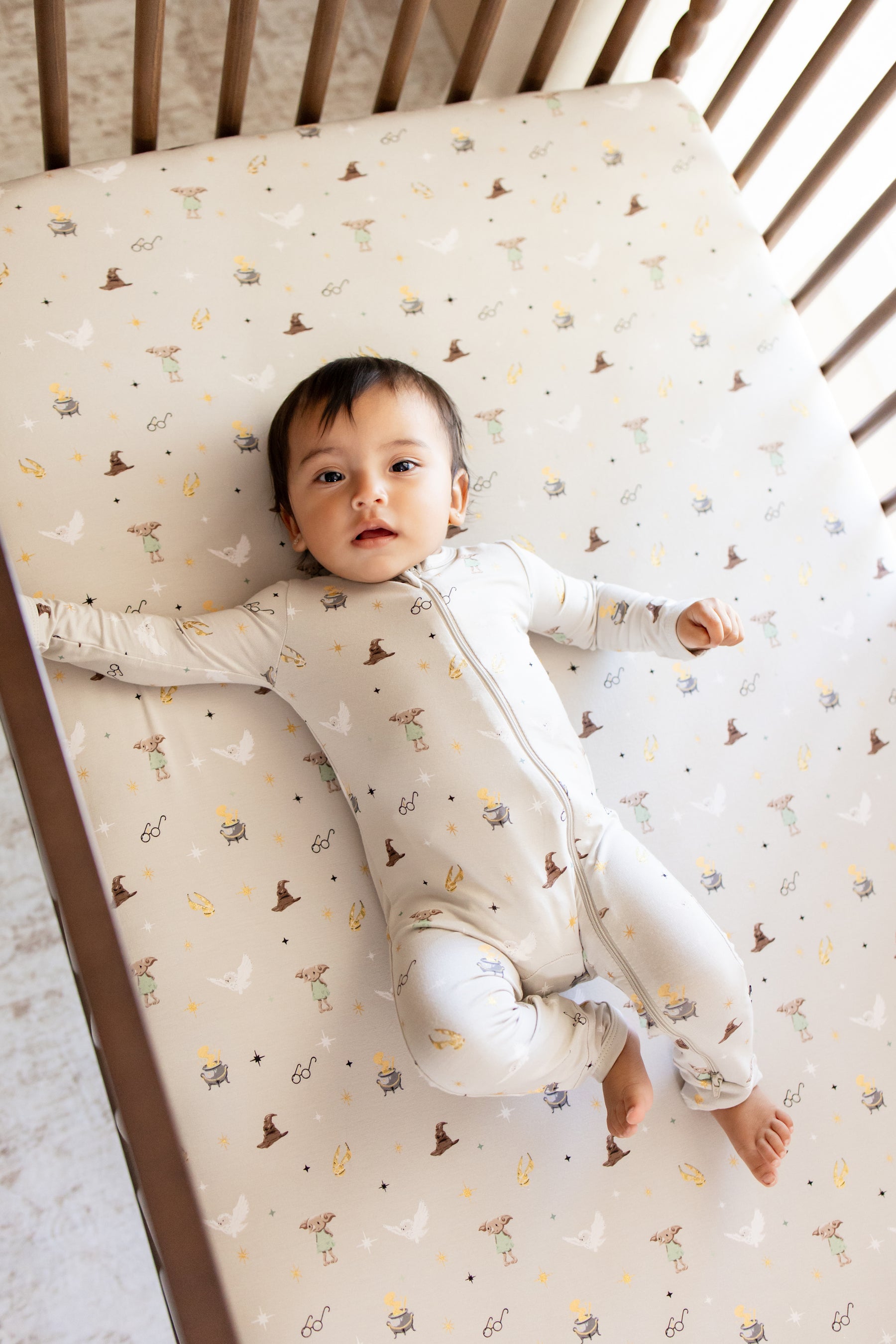 Baby wearing a zippered romper with Harry Potter prints lying in a crib with a similar print crib sheet.