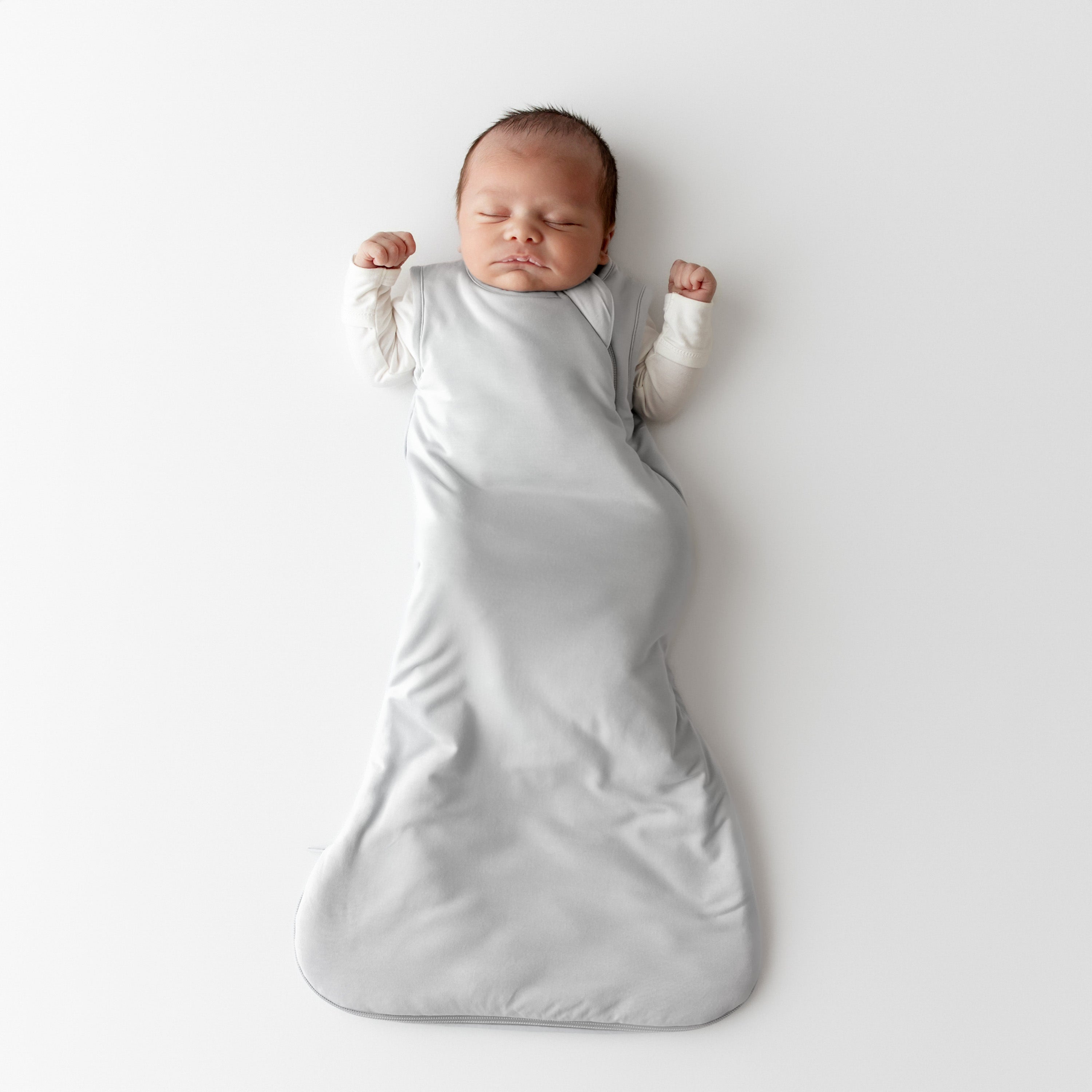 Newborn baby asleep in a light grey kyte baby sleep bag on a white background