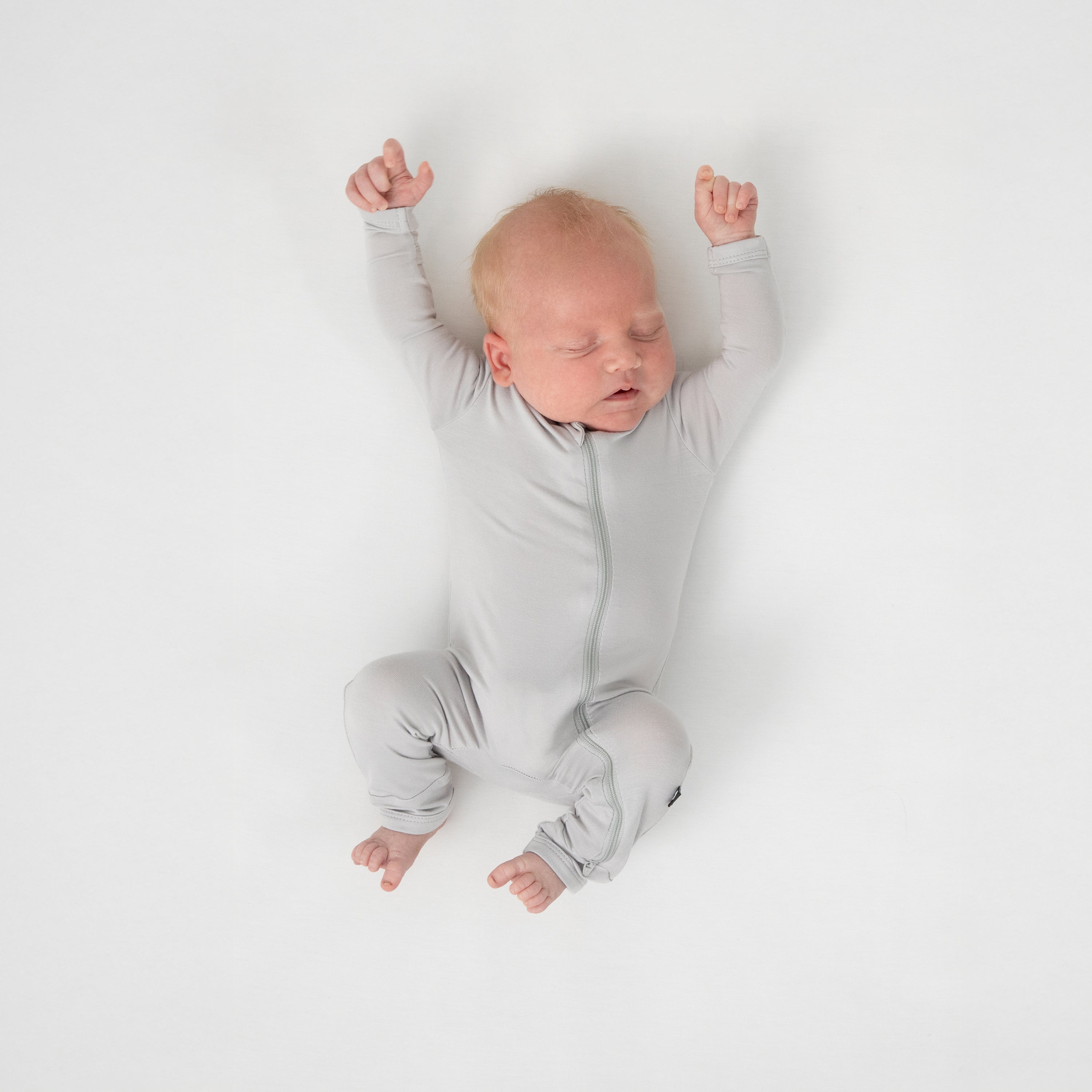 Sleeping newborn wearing the Zippered Footie in Storm laying on a white crib sheet