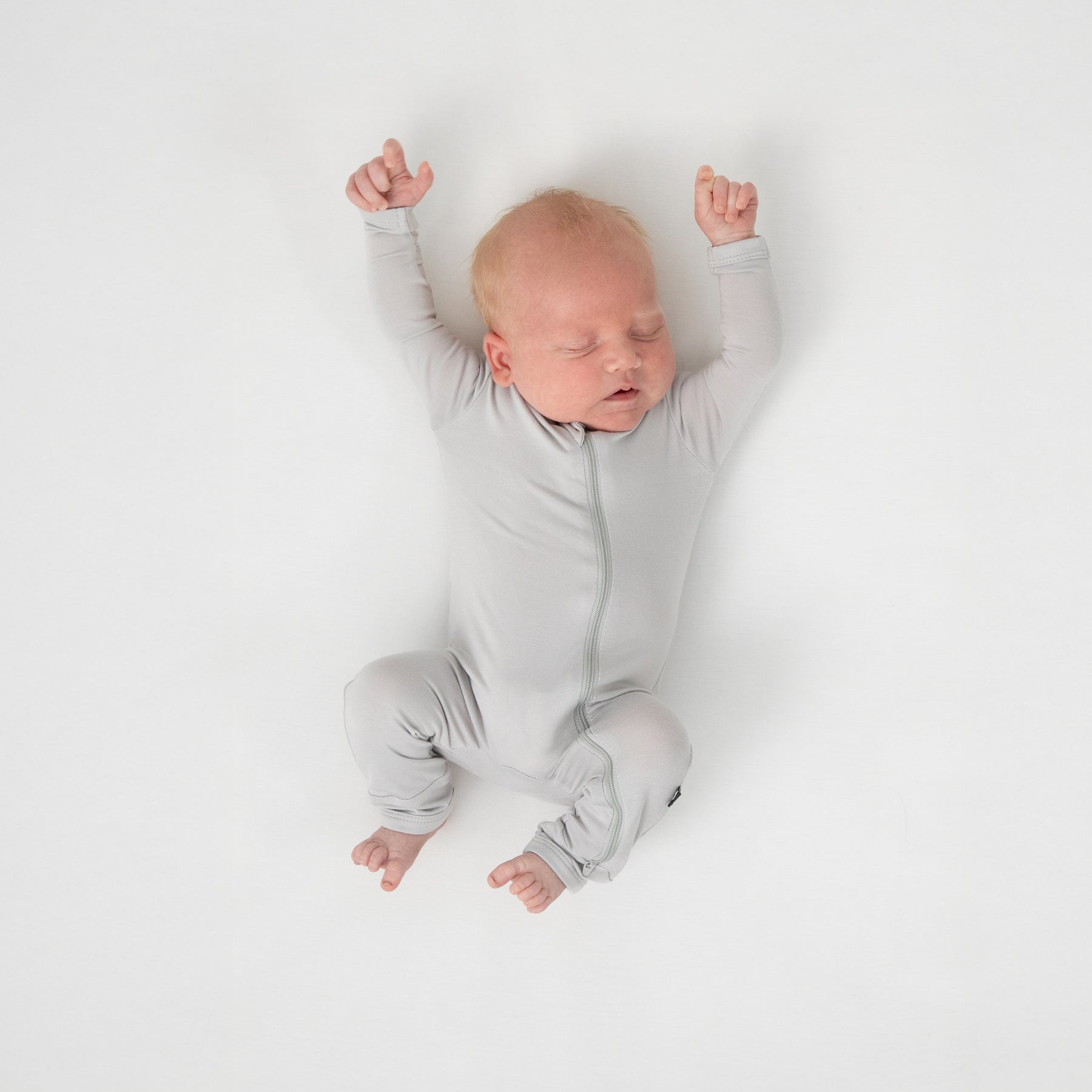 Sleeping newborn wearing the Zippered Footie in Storm laying on a white crib sheet