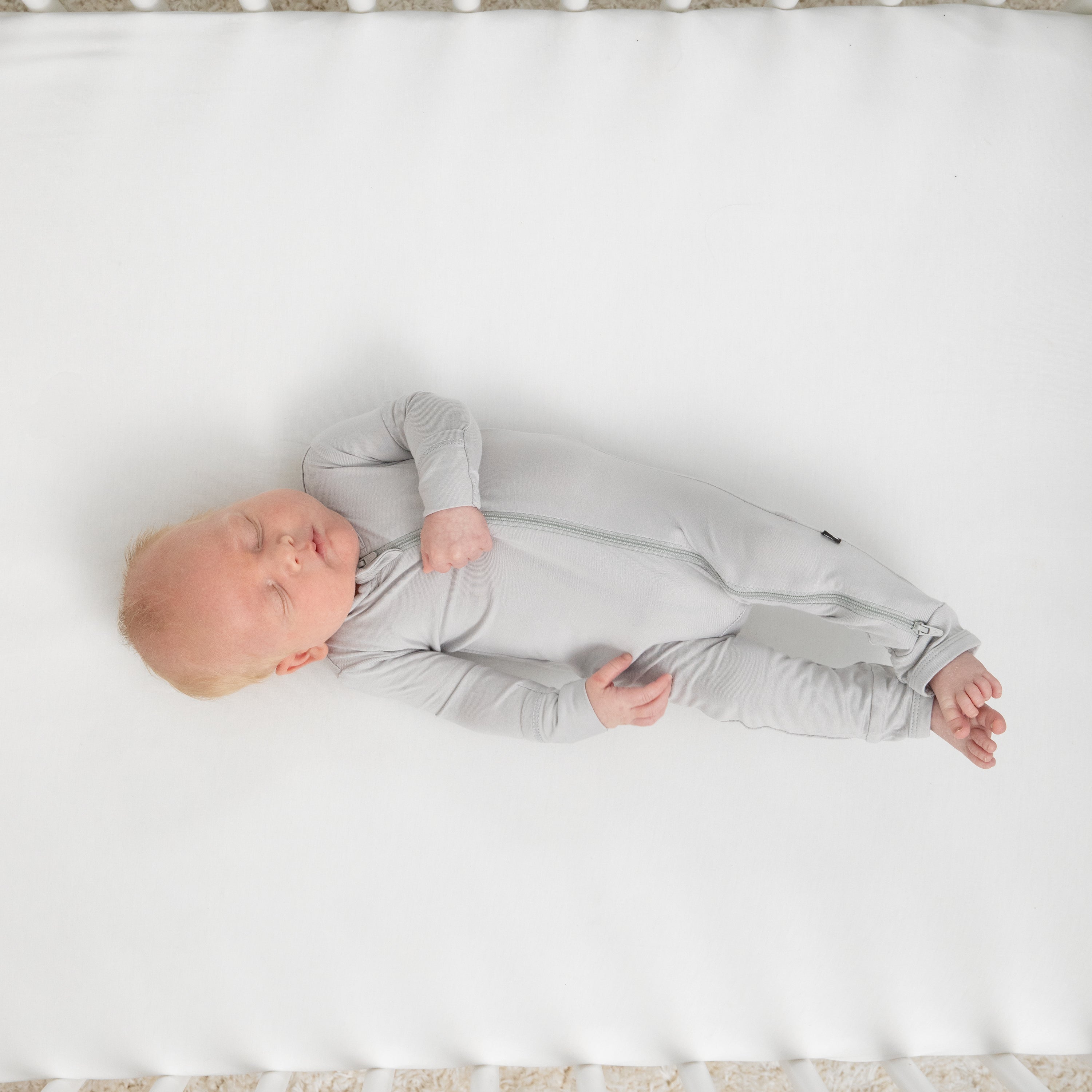 Infant sleeping in a crib on a white crib sheet wearing the Zippered Footie in Storm