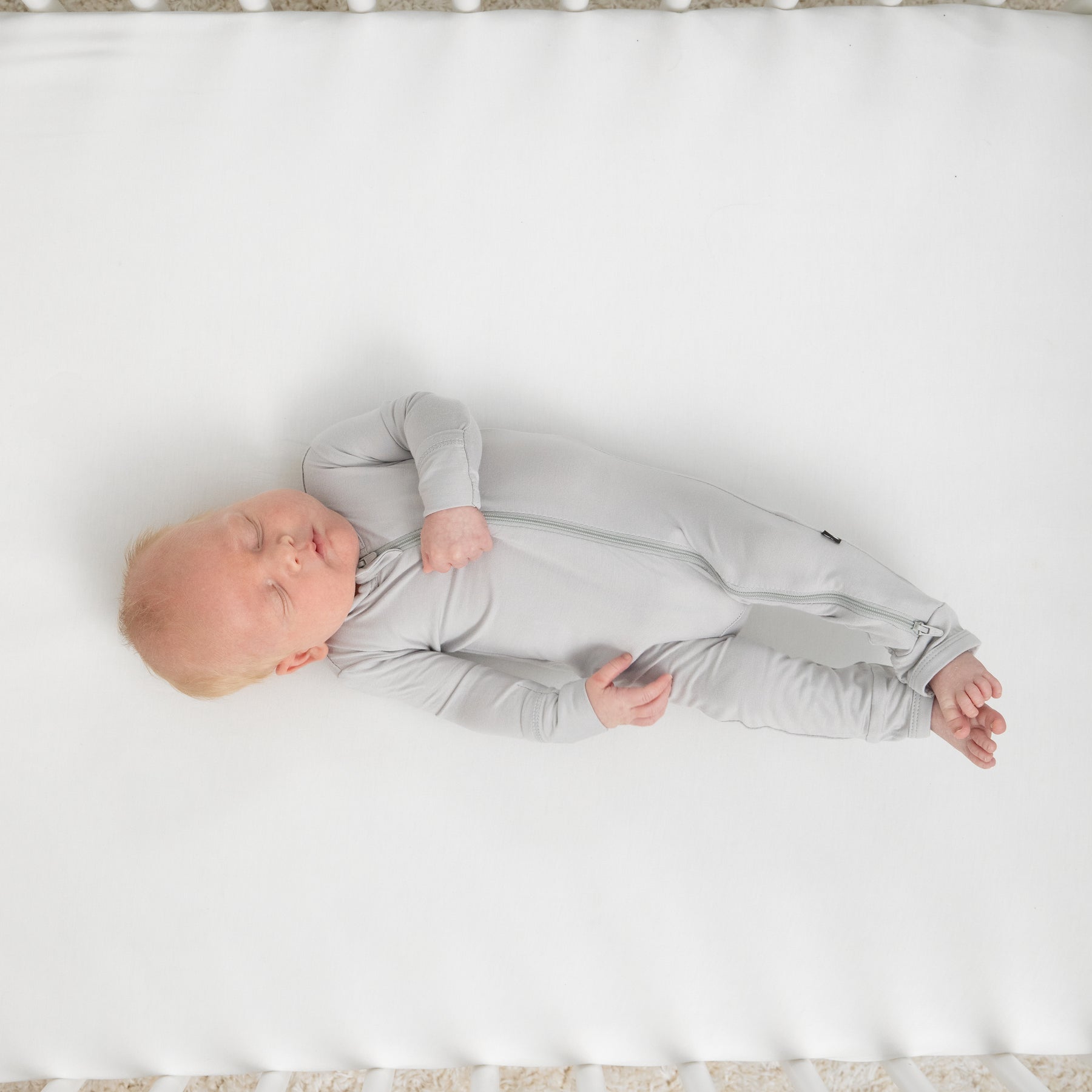 Infant sleeping in a crib on a white crib sheet wearing the Zippered Footie in Storm