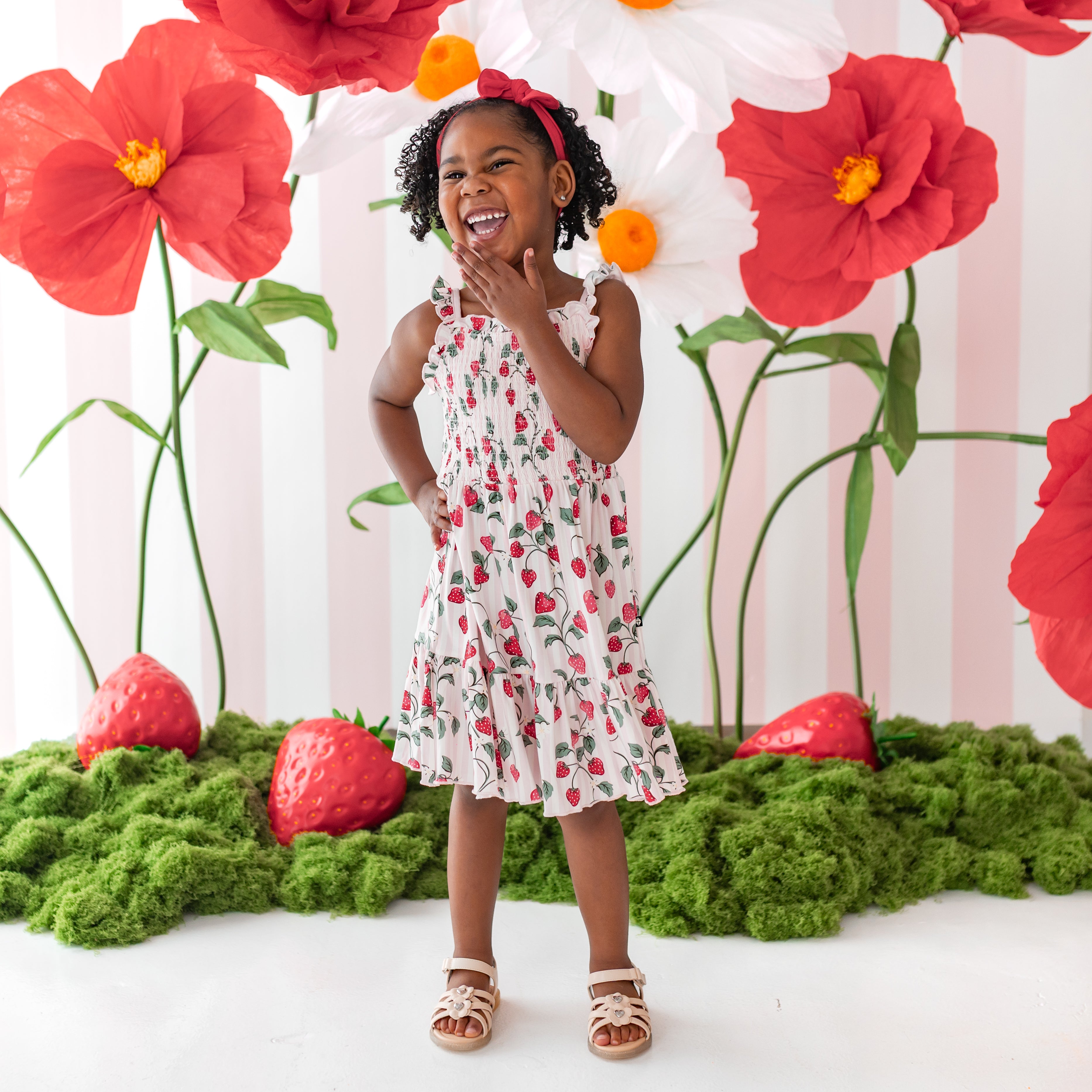 Laughing girl wearing the Smocked Dress in Strawberry Fields standing in front of a strawberry themed backdrop