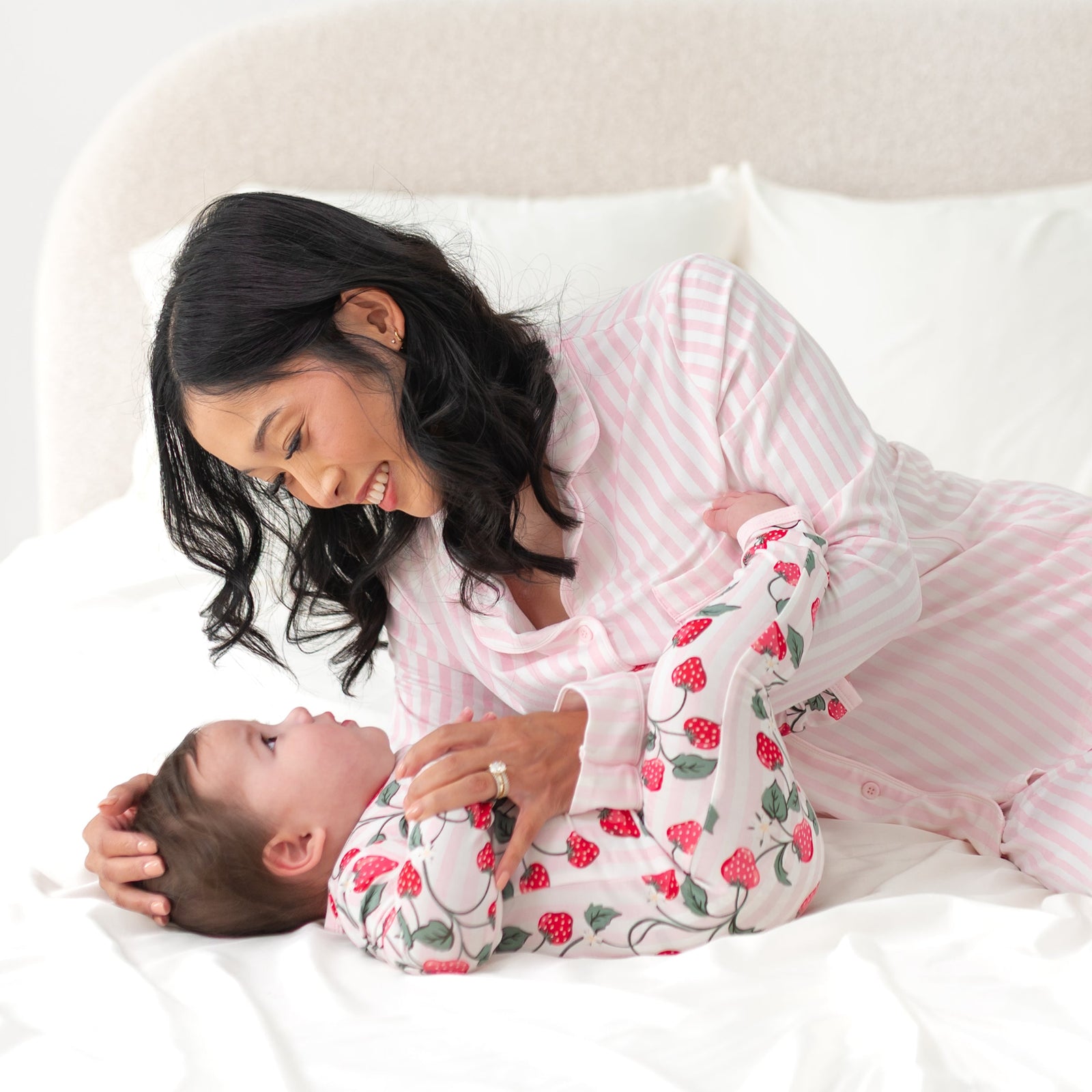 Mom and baby snuggled on a white bed wearing pink stripes and strawberry pajamas