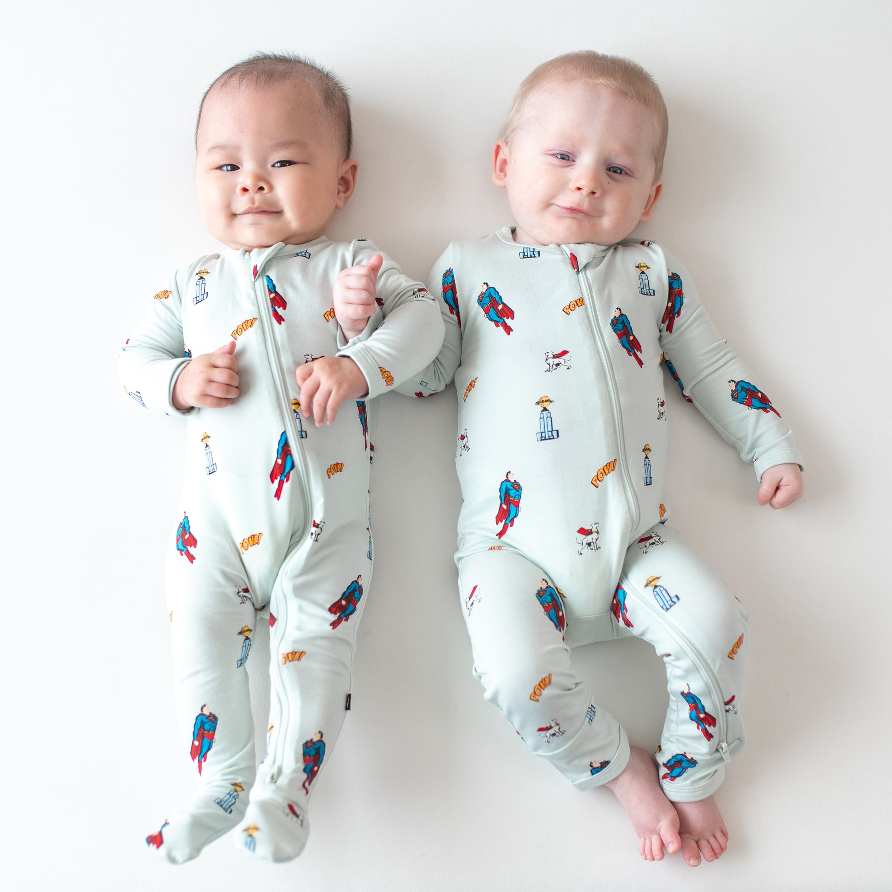 Two infants laying side by side wearing the Zippered Romper and Zippered Footie in Superman™