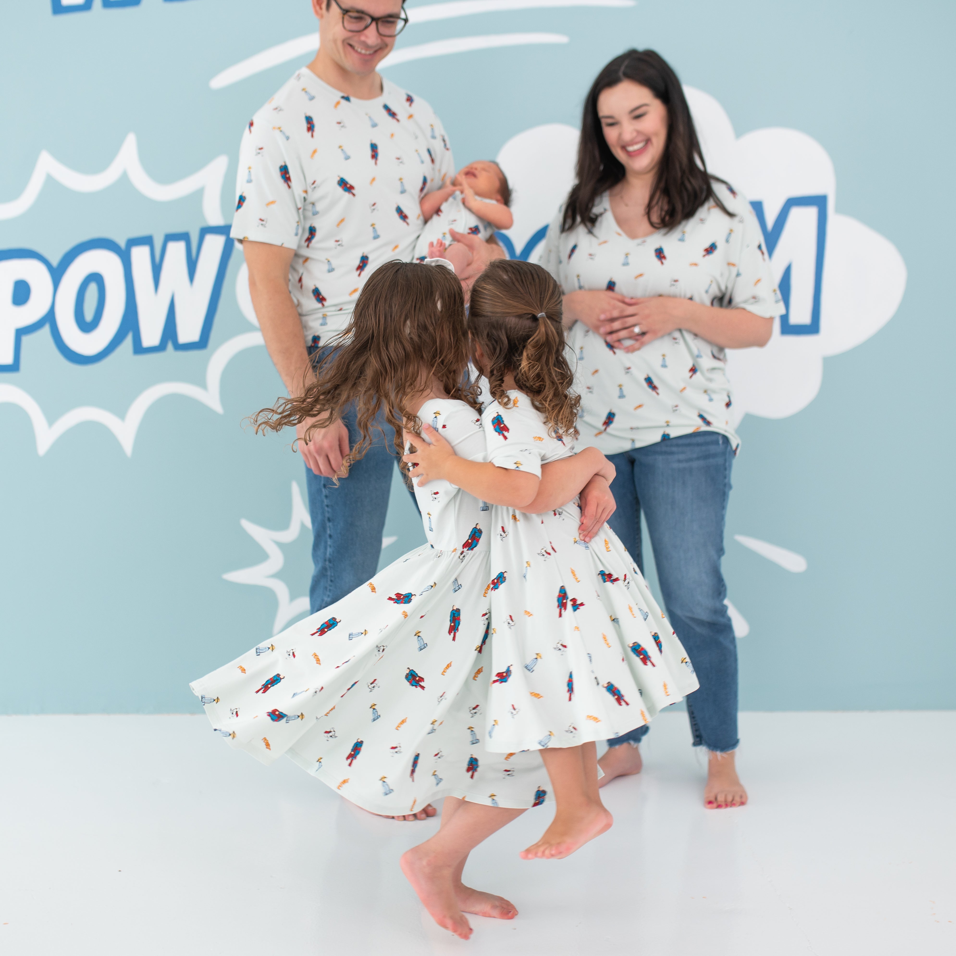 Big sister twirling younger sister around both wearing the Twirl Dress in Superman™ in front of their parents and younger sibling who are also wearing items from the Superman™ collection