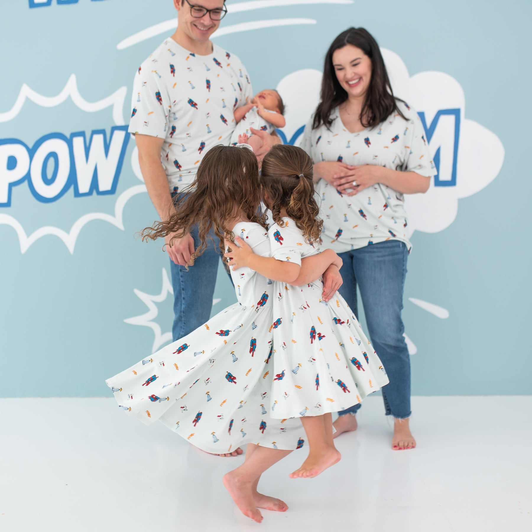 Big sister twirling younger sister around both wearing the Twirl Dress in Superman™ in front of their parents and younger sibling who are also wearing items from the Superman™ collection