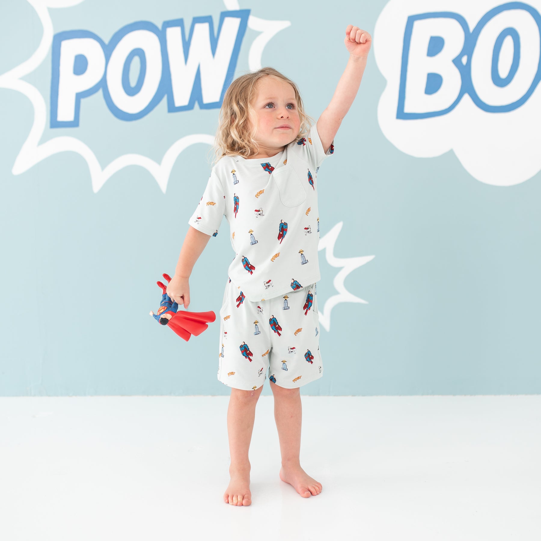 Toddler standing with one hand in the air and the other holding a superman action figure wearing the Toddler Crew Neck Tee in Superman™ with matching drawstring shorts