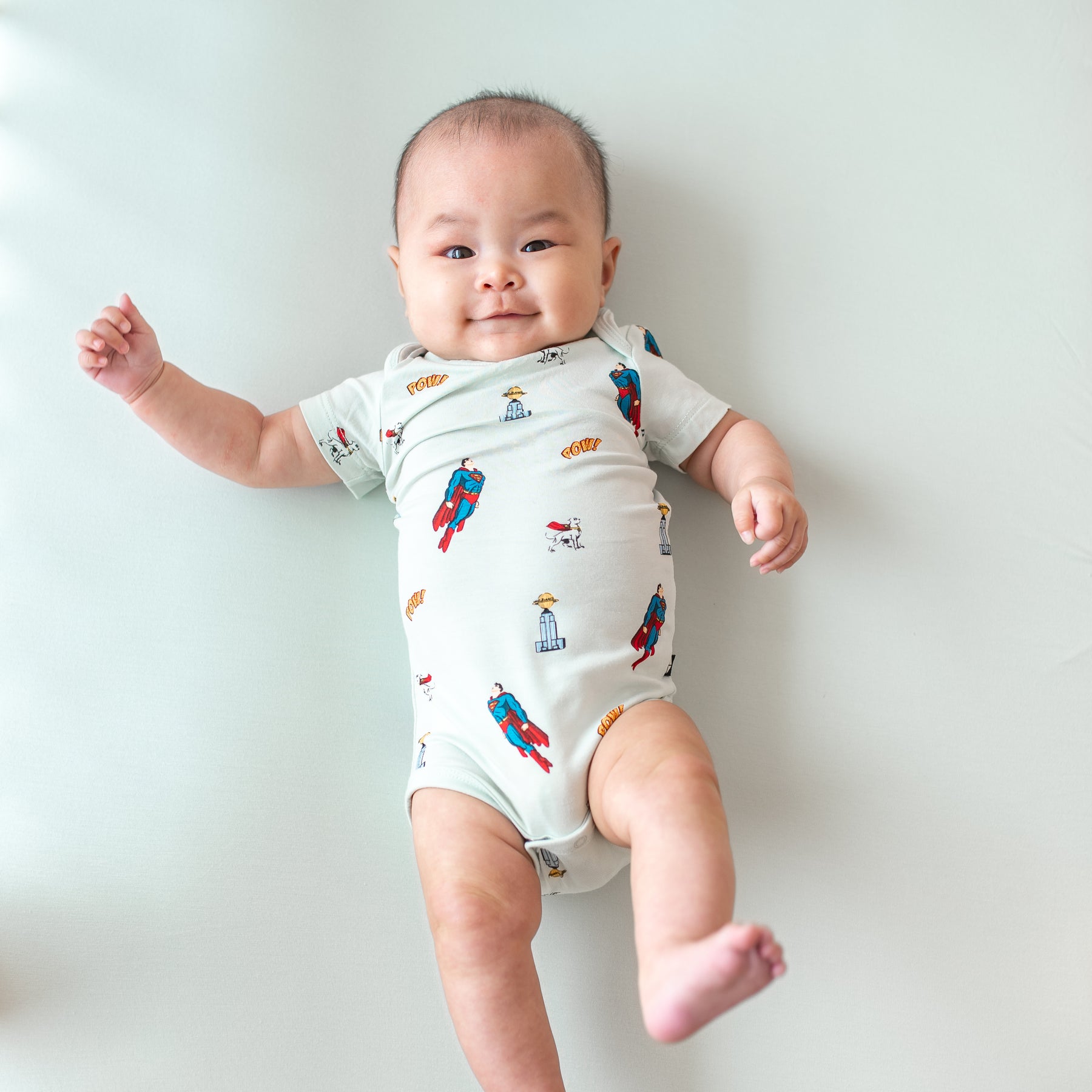 Infant model laying down wearing the Bodysuit in Superman™ with one leg up in the air
