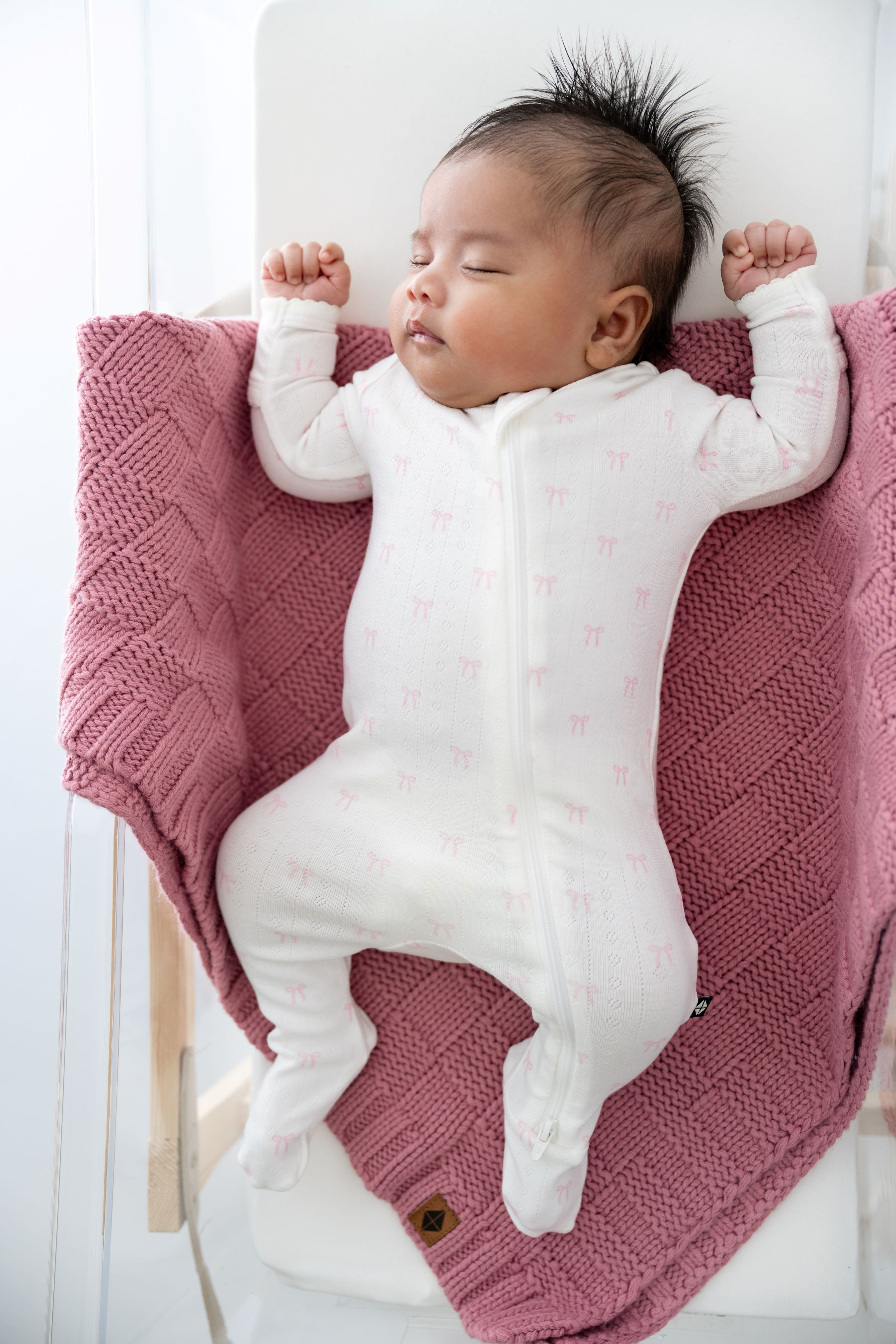 Sleeping newborn wearing the Pointelle Zippered Footie in Sweet Pink Bow laying on a dusty rose basket weave blanket