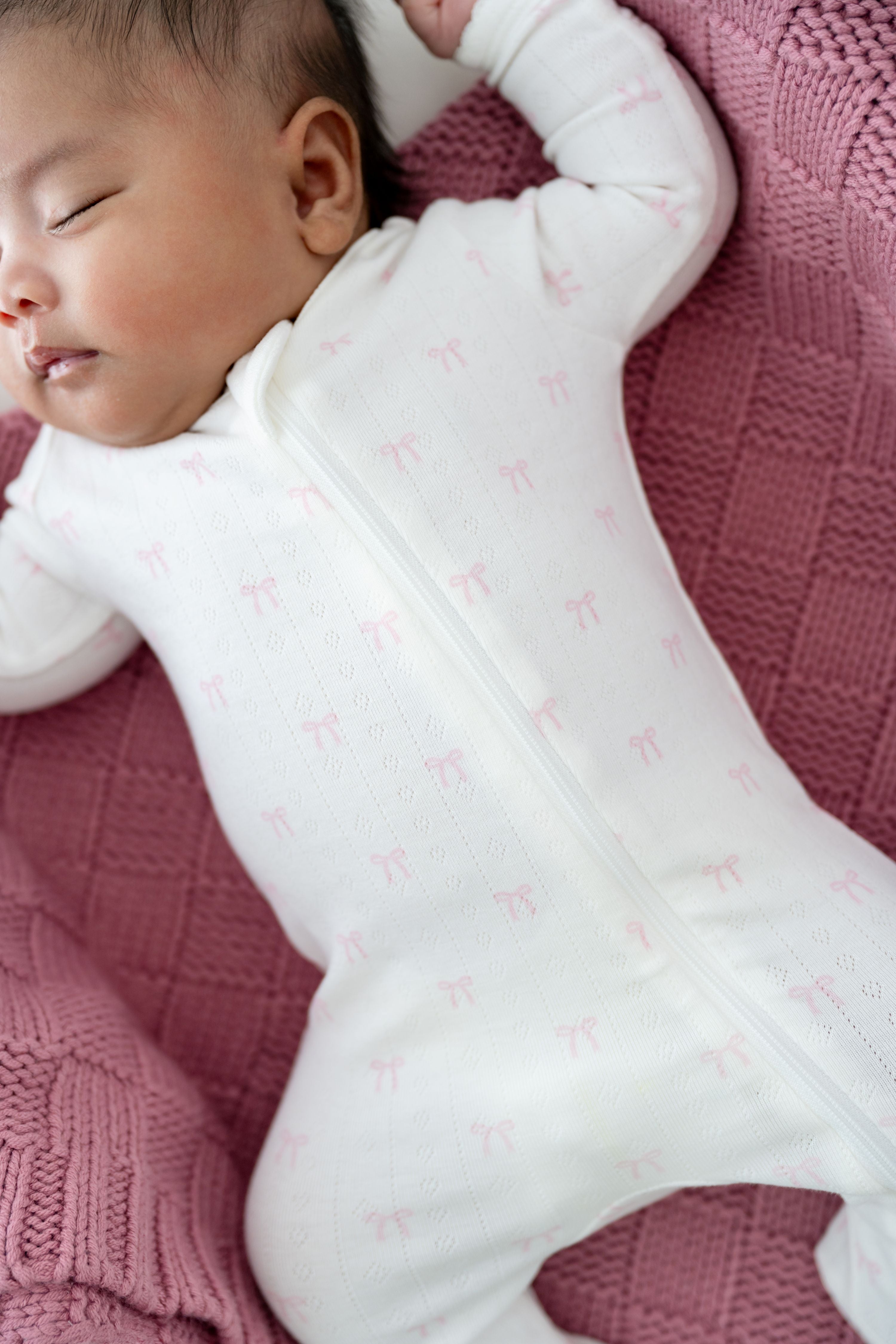 Close up of sleeping baby wearing the Pointelle Zippered Footie in Sweet Pink Bow on a dusty rose colored blanket
