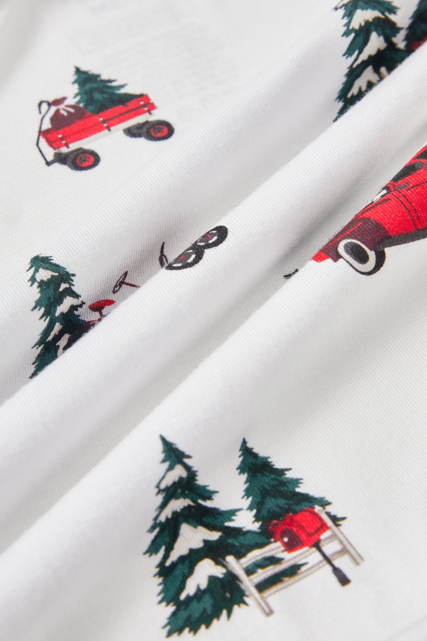 White fabric with a repeating print of red wagons, green pine trees, and a red sled, shown in a close-up folded view.