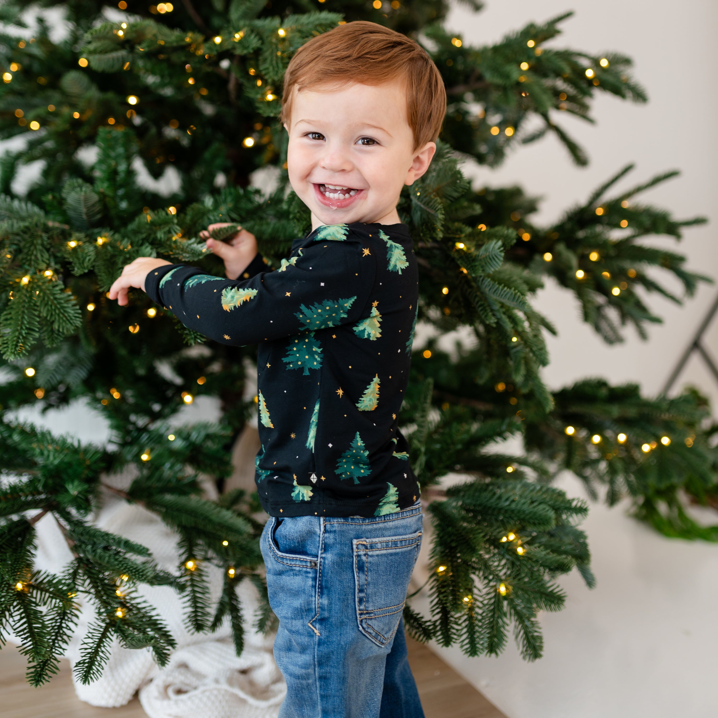 Young bot holding a branch on a Christmas tree wearing the Long Sleeve Toddler Crew Neck Tee in Twinkle Tree paired with medium wash jeans