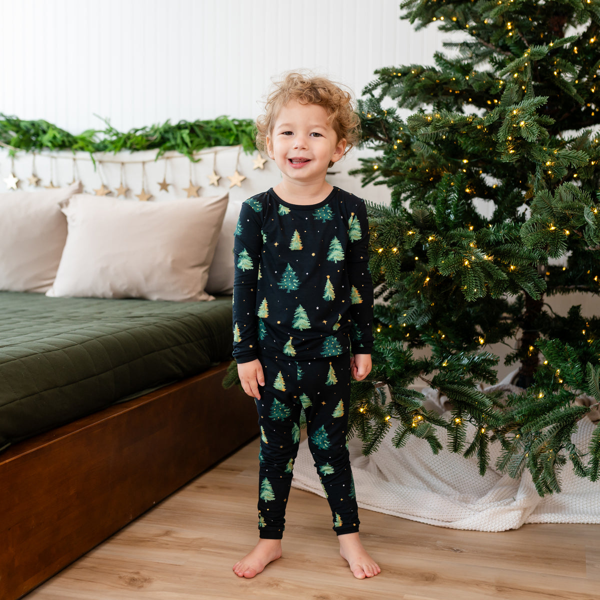 Smiling young boy wearing the Long Sleeve Pajamas in Twinkle Tree standing between a christmas tree and bed