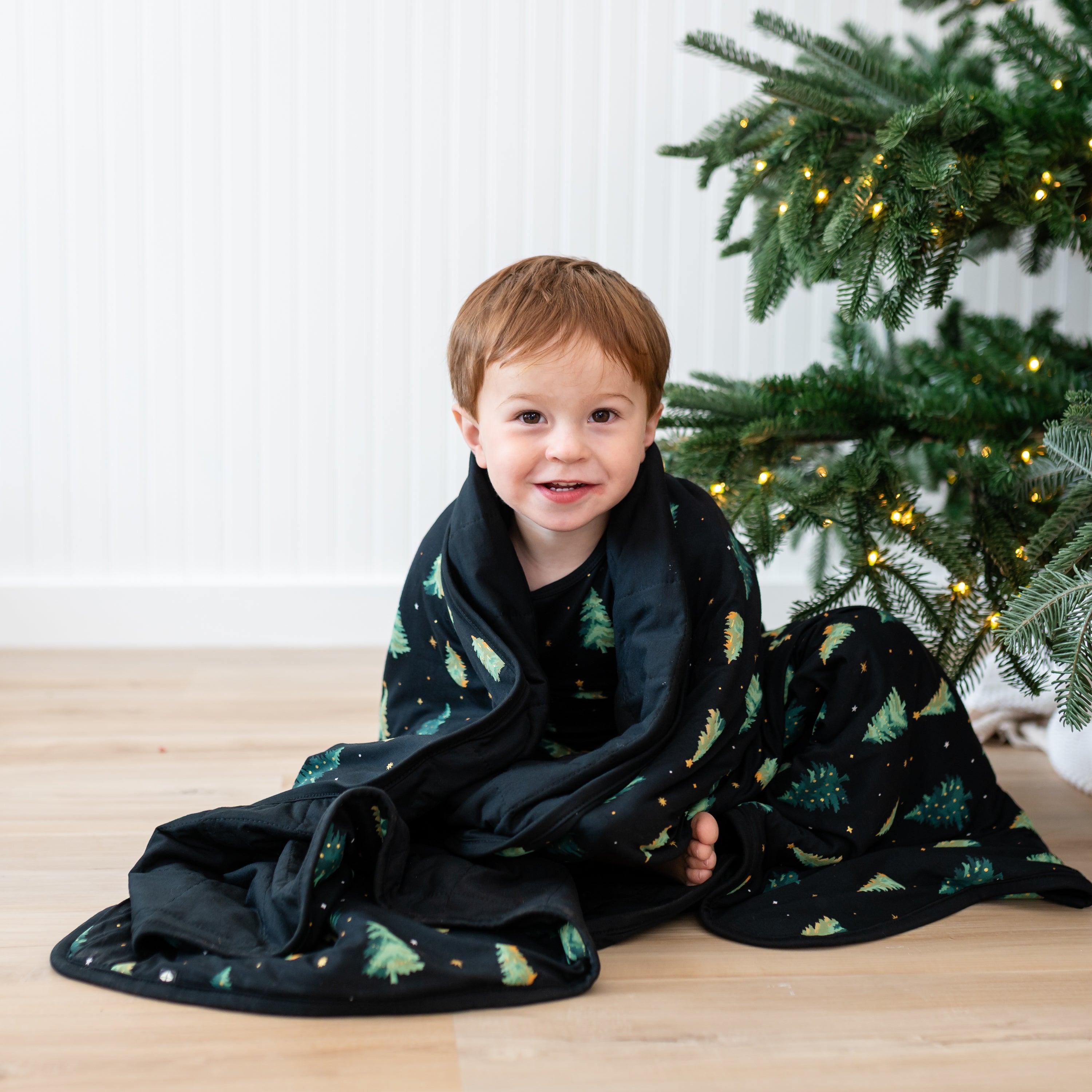 Toddler sitting on the floor with the Toddler Blanket in Twinkle Tree 1.0 wrapped around himself