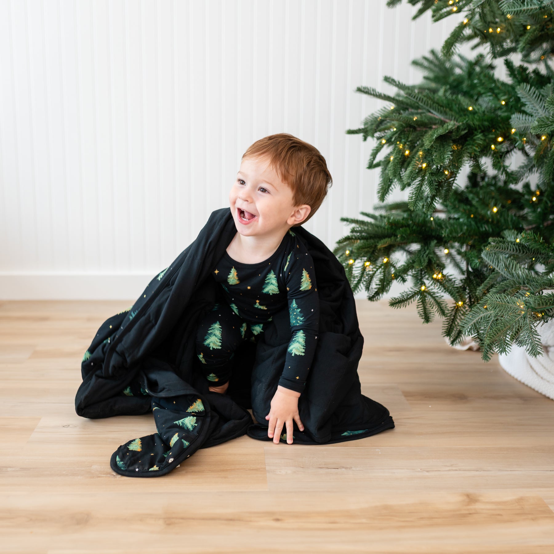 Toddler sitting on the floor wearing the Long sleeve toddler pajama set in Twinkle tree with the Toddler Blanket in Twinkle Tree 1.0 wrapped around him