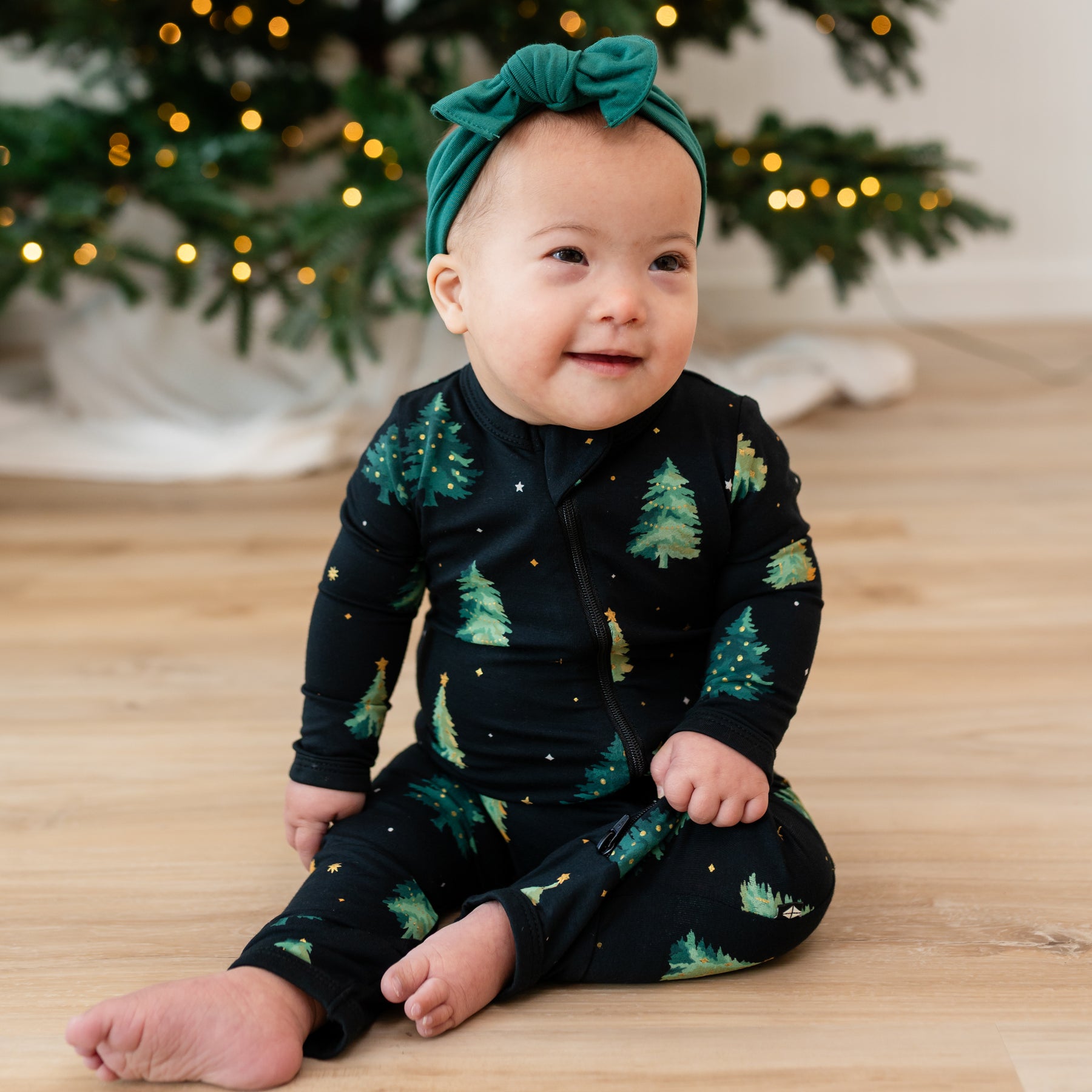 infant sitting wearing the Zippered Romper in Twinkle Tree and emerald knotted bow headband