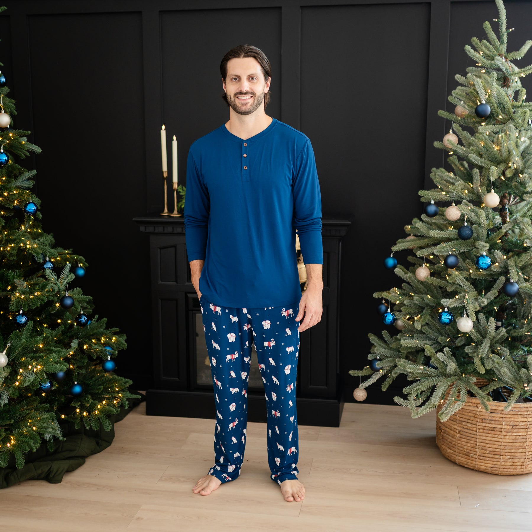 Smiling male model standing between two decorated christmas trees wearing the Men's Long Sleeve Henley Top in Tahoe and the Men's Lounge Pant in Frosty Friends