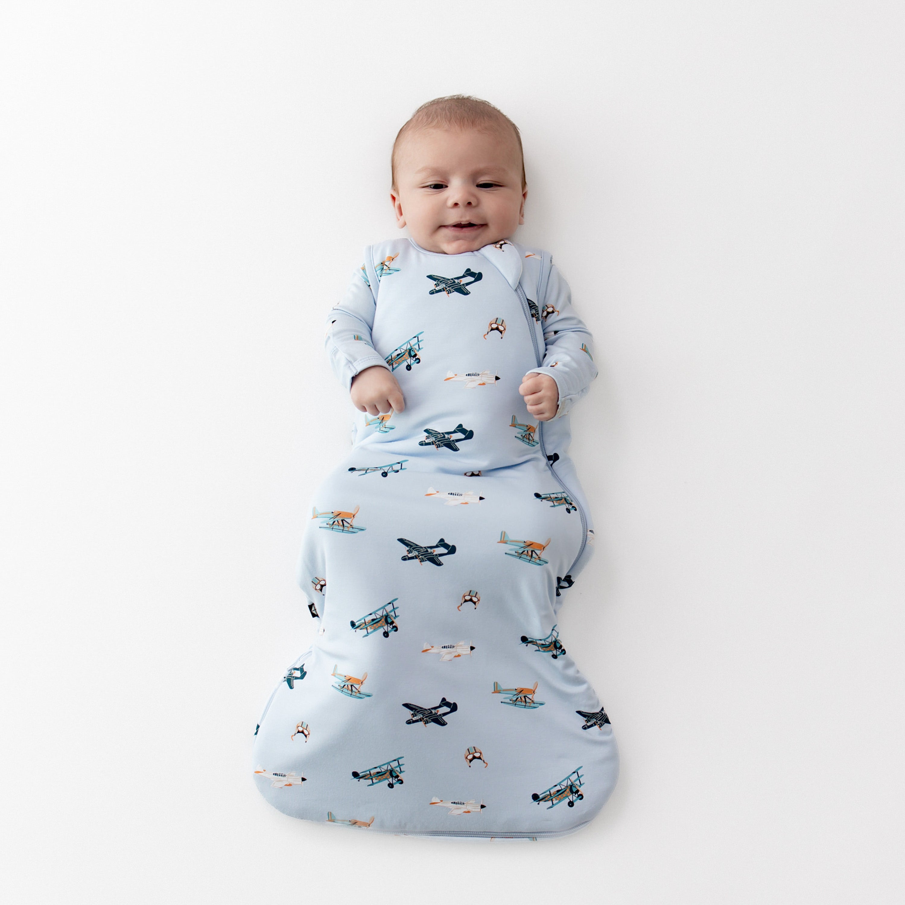 Infant laying on a white surface wearing the Sleep Bag in Take Off 1.0 with matching long sleeve romper on underneath
