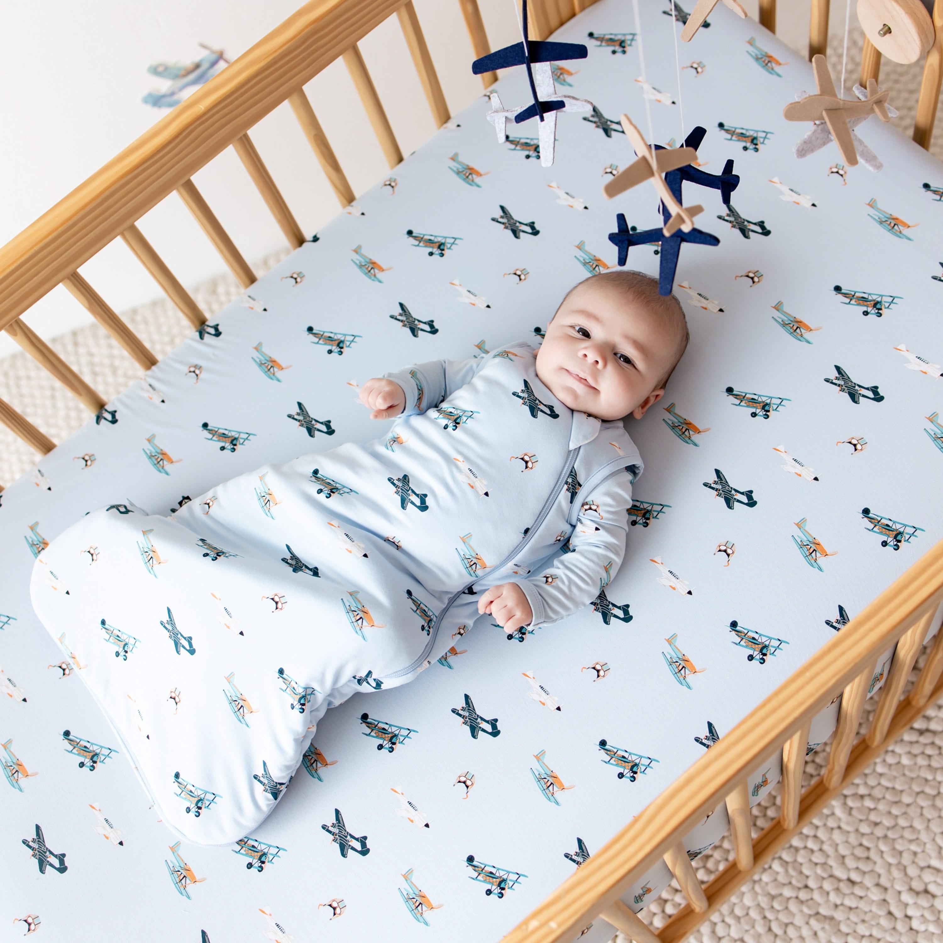 Infant laying in a crib on top of Take Off crib sheets wearing the Sleep Bag in Take Off 1.0 o