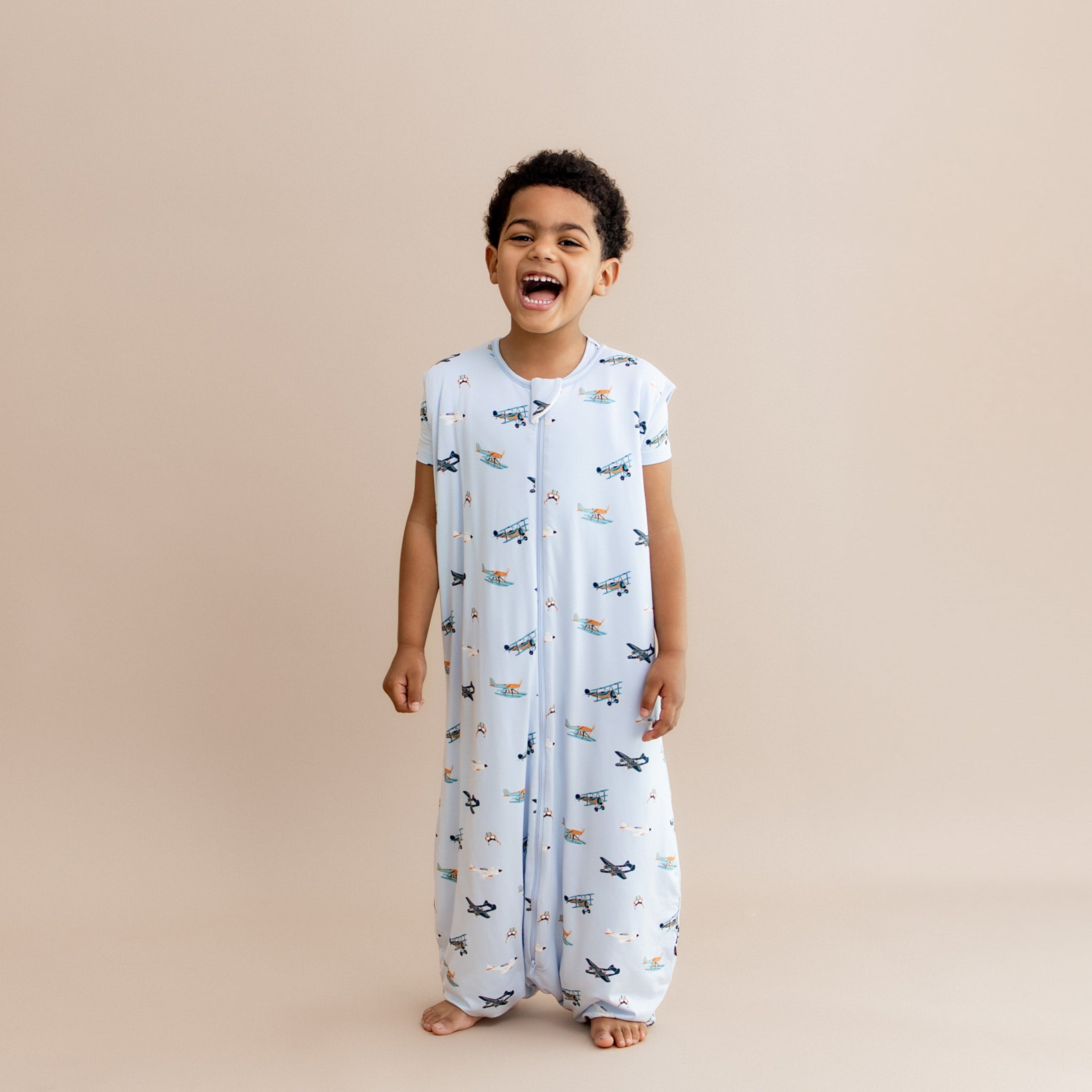 Laughing boy standing wearing the Sleep Bag Walker in Take Off 1.0 with matching short sleeve toddler pajamas on underneath