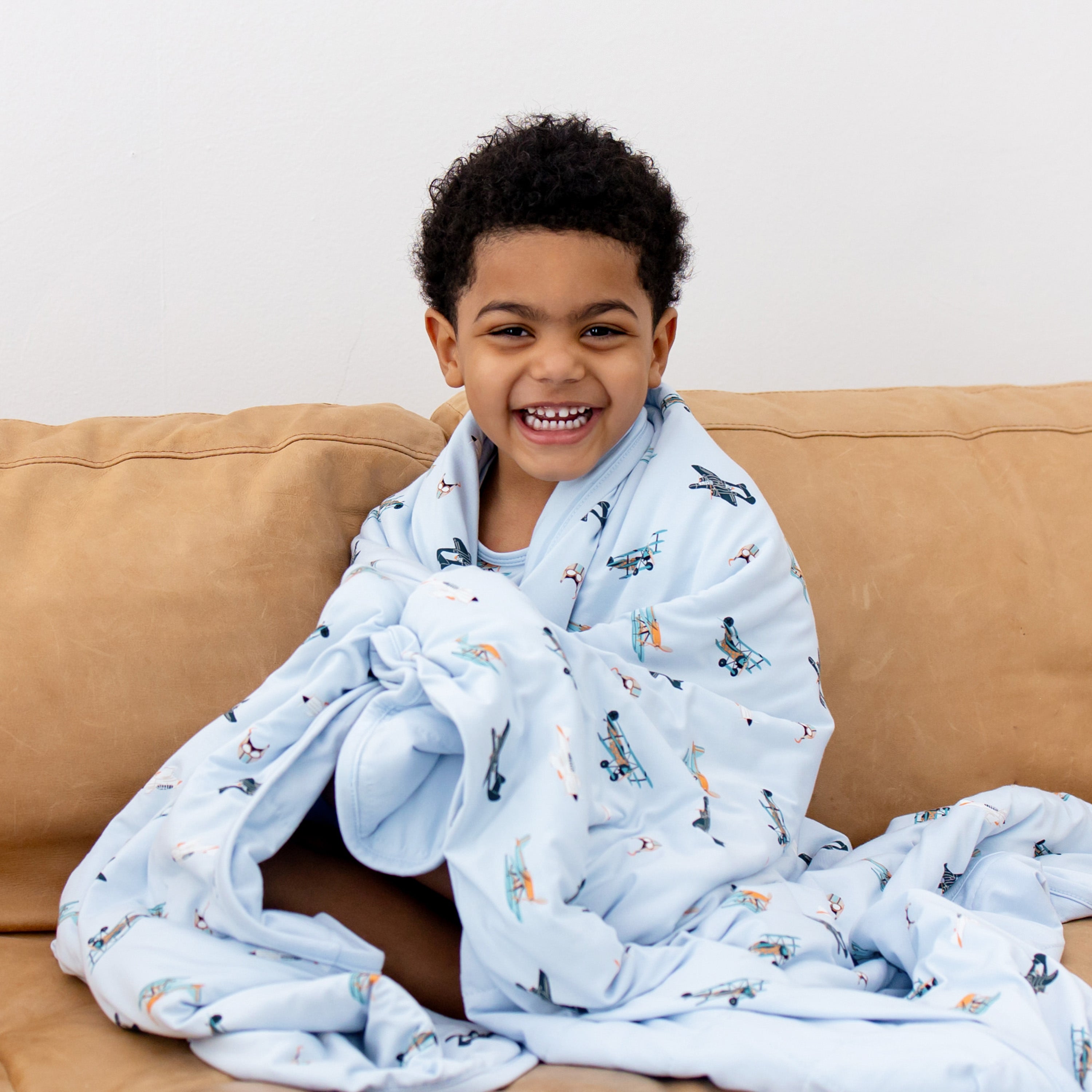 Close up of smiling boy sitting on a couch with the Toddler Blanket in Take Off 1.0 around his shoulders