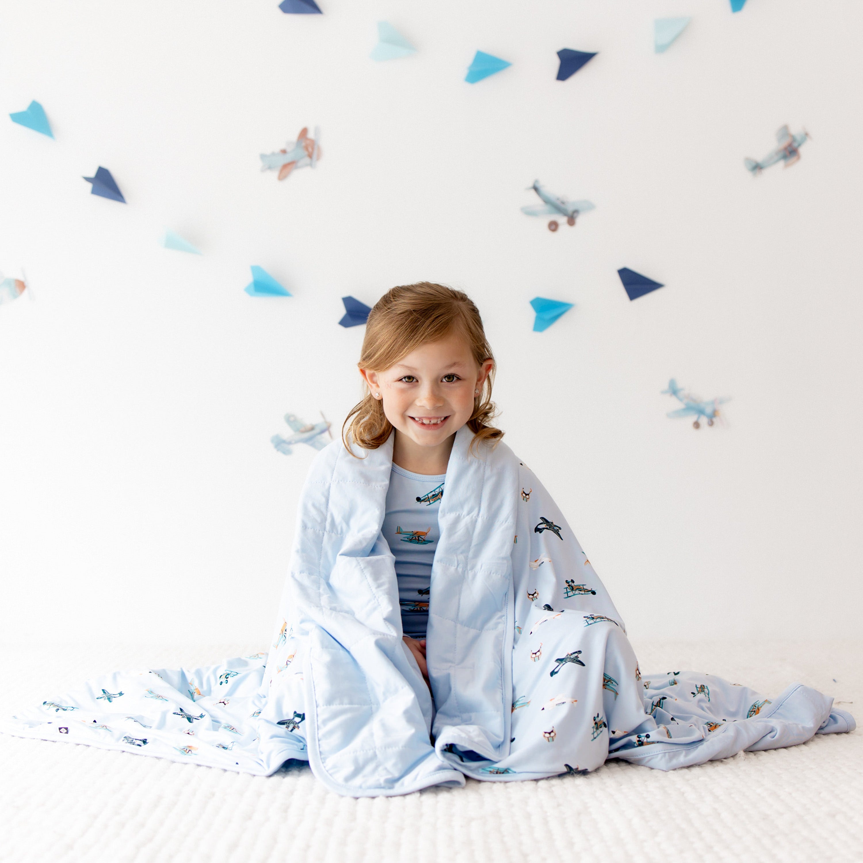 Young girl sitting on a white carpet with the Youth Blanket in Take Off 1.0 around her shoulders