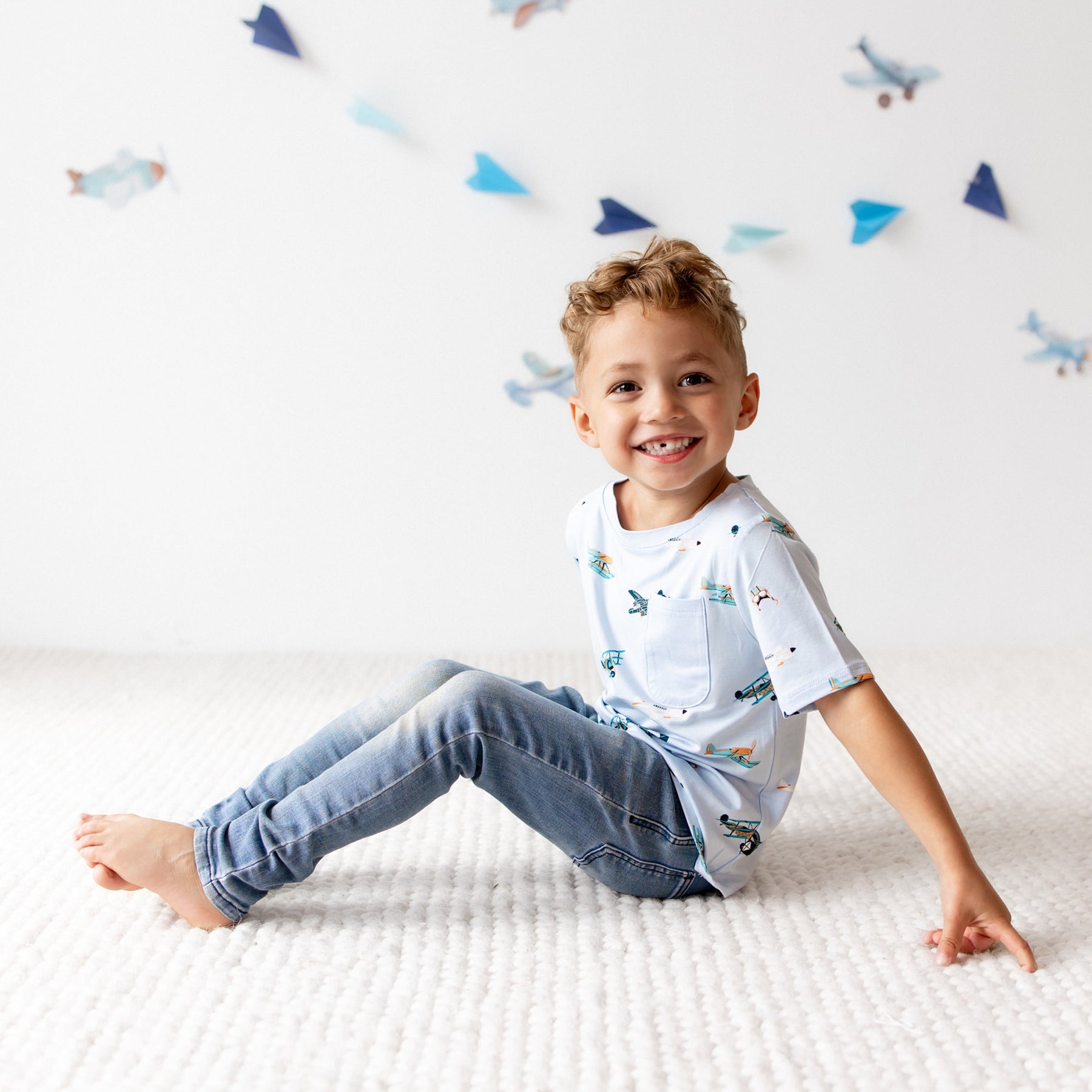 Young boy sitting on a cream floor wearing the Toddler Crew Neck Tee in Take Off with light wash jeans
