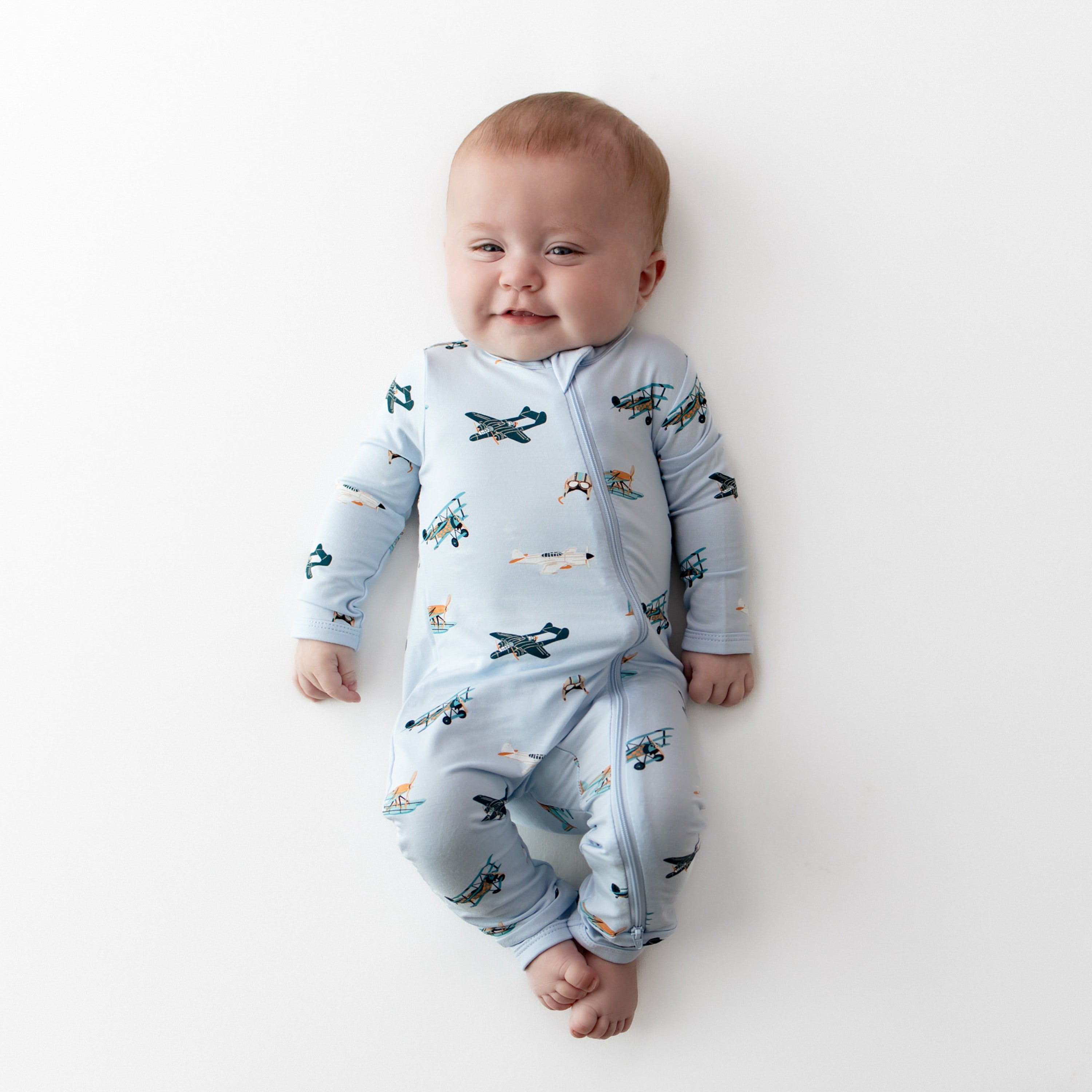 Smiling infant laying on a white surface wearing the Zippered Romper in Take Off