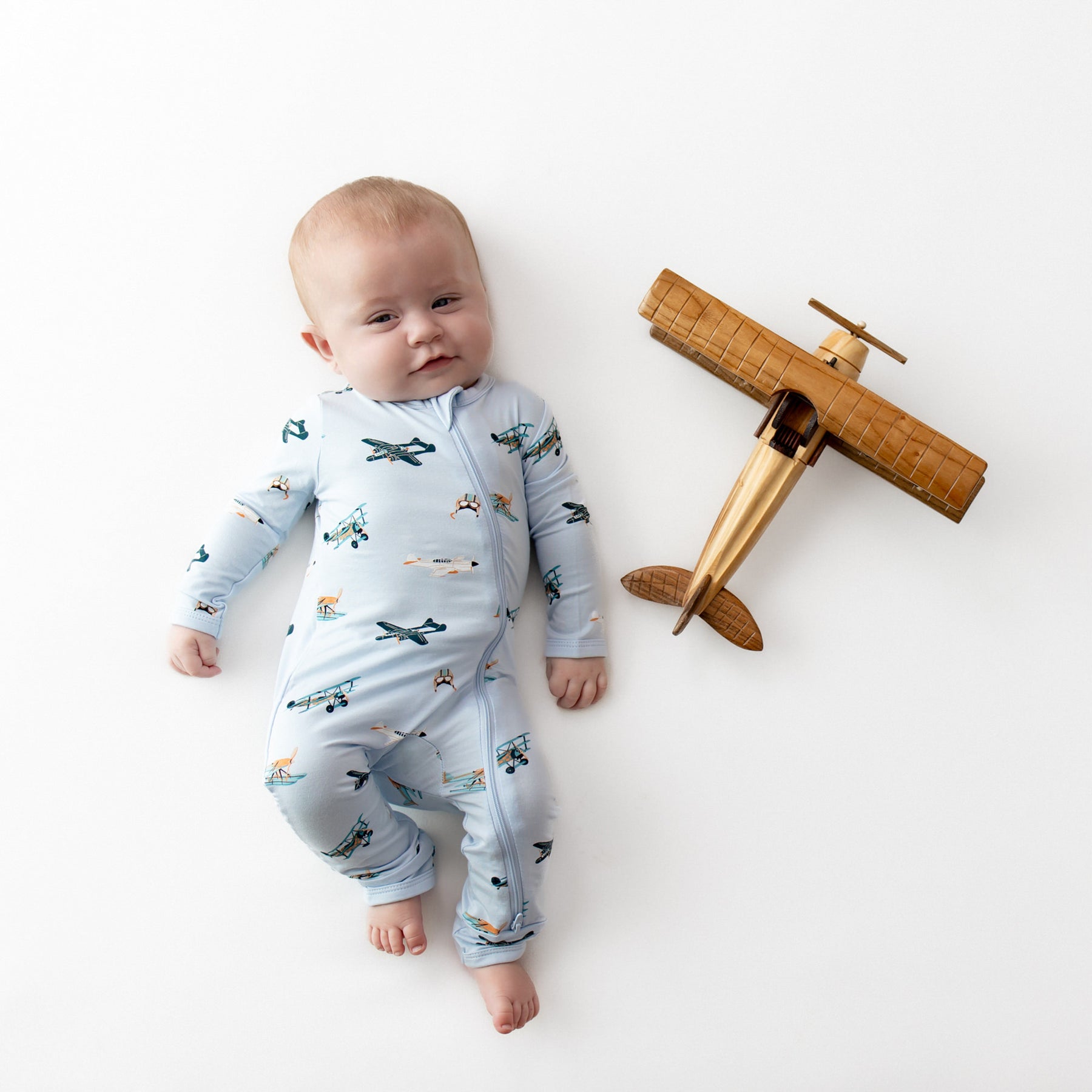 Infant laying beside a wooden toy plane wearing the Zippered Romper in Take Off