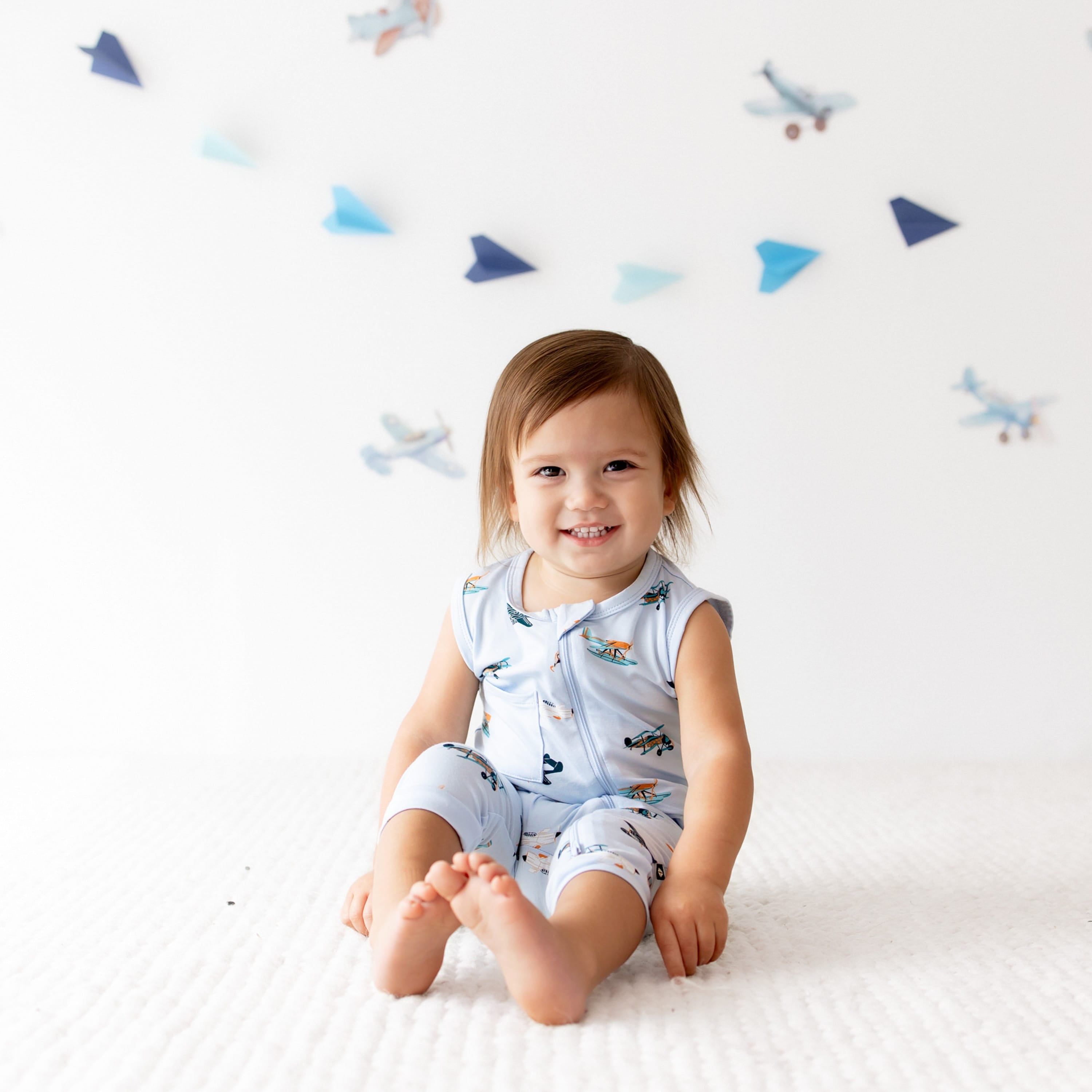 Young toddler sitting on a white carpet wearing the Zippered Sleeveless Romper in Take Off