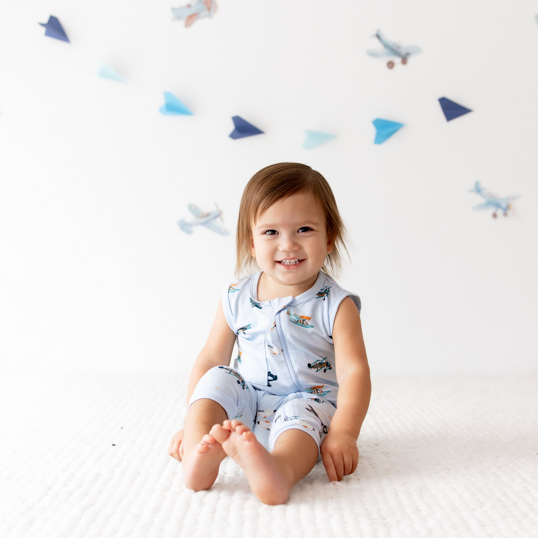 Young toddler sitting on a white carpet wearing the Zippered Sleeveless Romper in Take Off