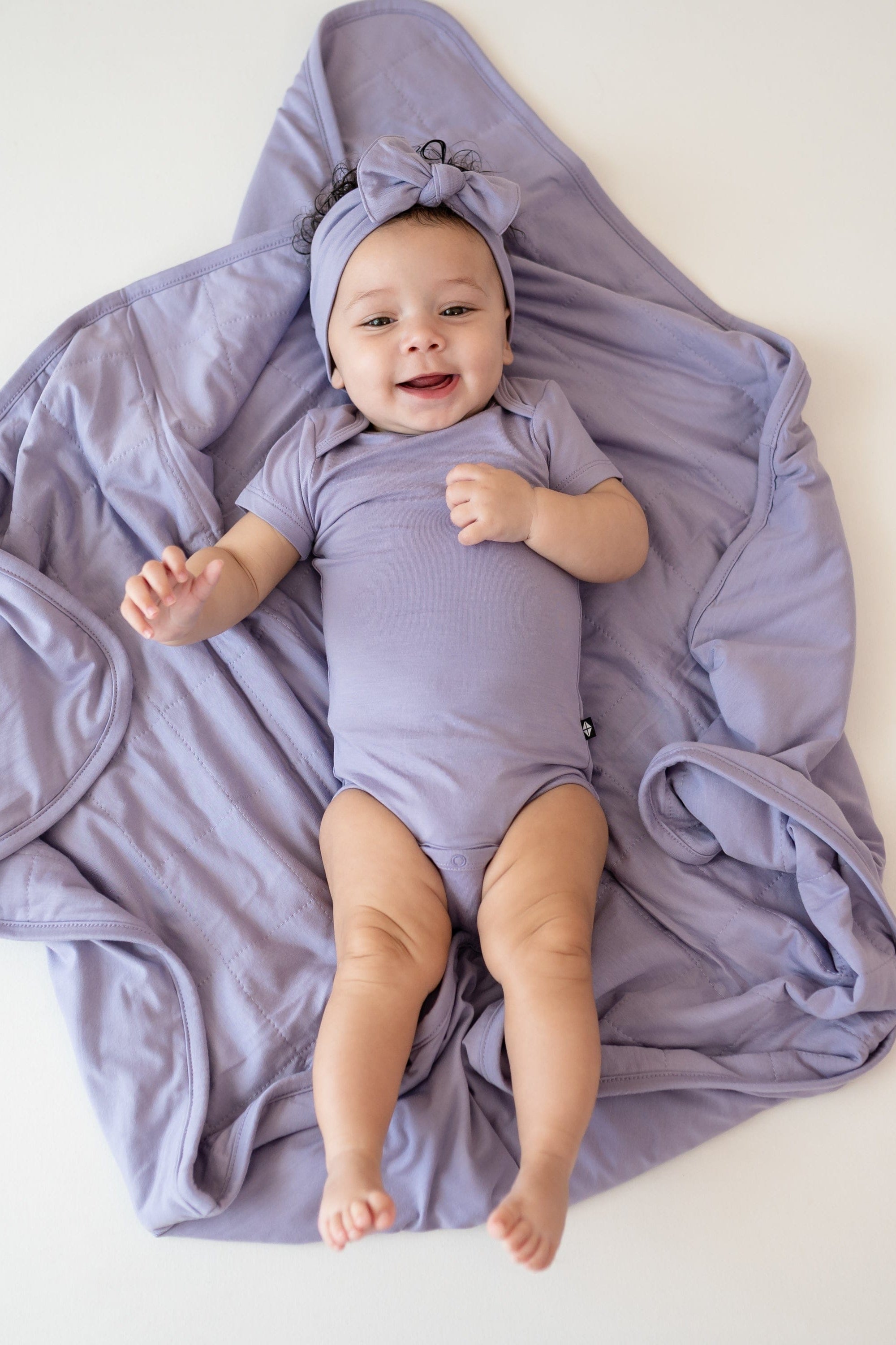 Baby laying on Baby Blanket in Taro 1.0 wearing bodysuit and bow in taro