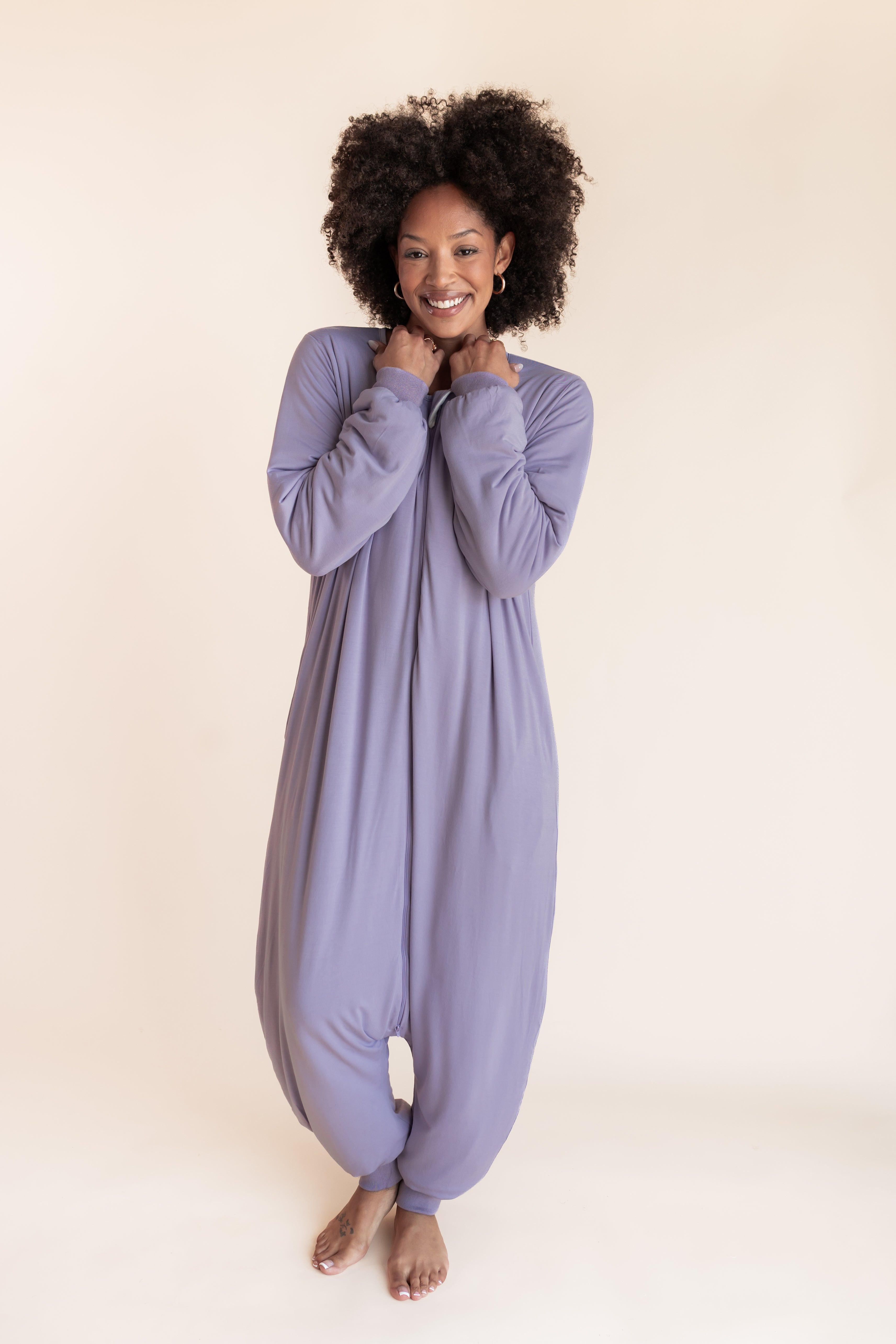A woman with curly black hair wearing a lavender-purple long-sleeve harem-style sleep sack, standing against a solid light beige background.