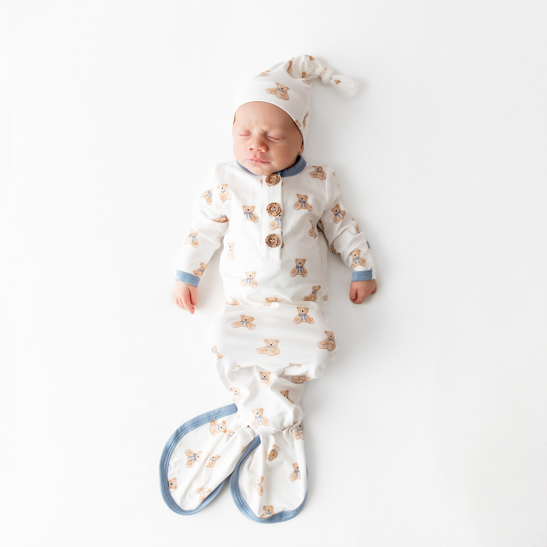 Sleeping newborn laying on a white surface wearing the Knotted Gown with Hat Set in Teddy Bear 