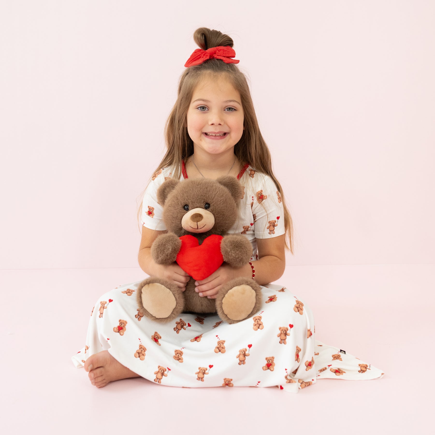 Young girl wearing the Twirl Dress in Bear Hearts sitting on the floor in front of a pink background holding Kyte Baby's Valentine's Teddy Bear
