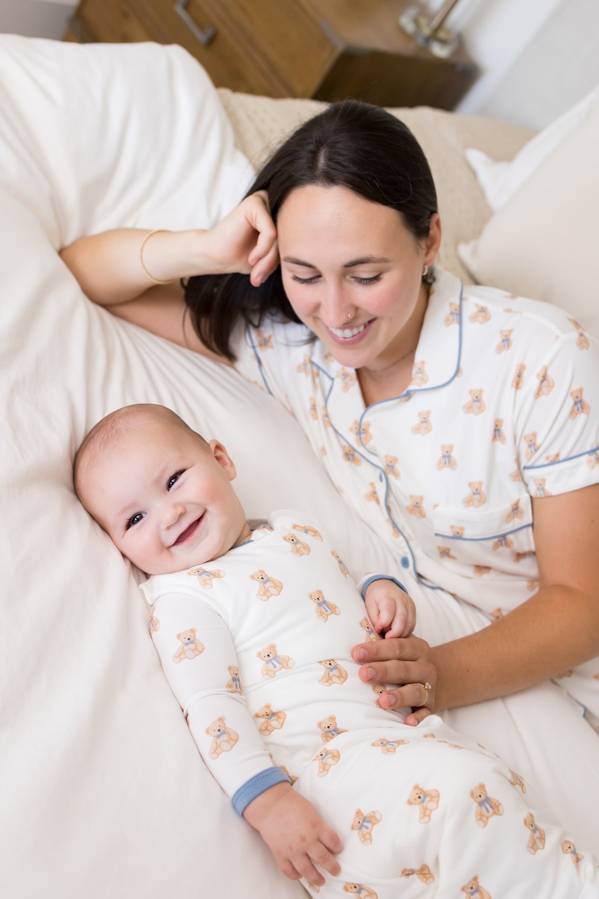 A smiling woman and baby lying on a bed, both wearing matching white pajamas with a teddy bear print and blue piping details.