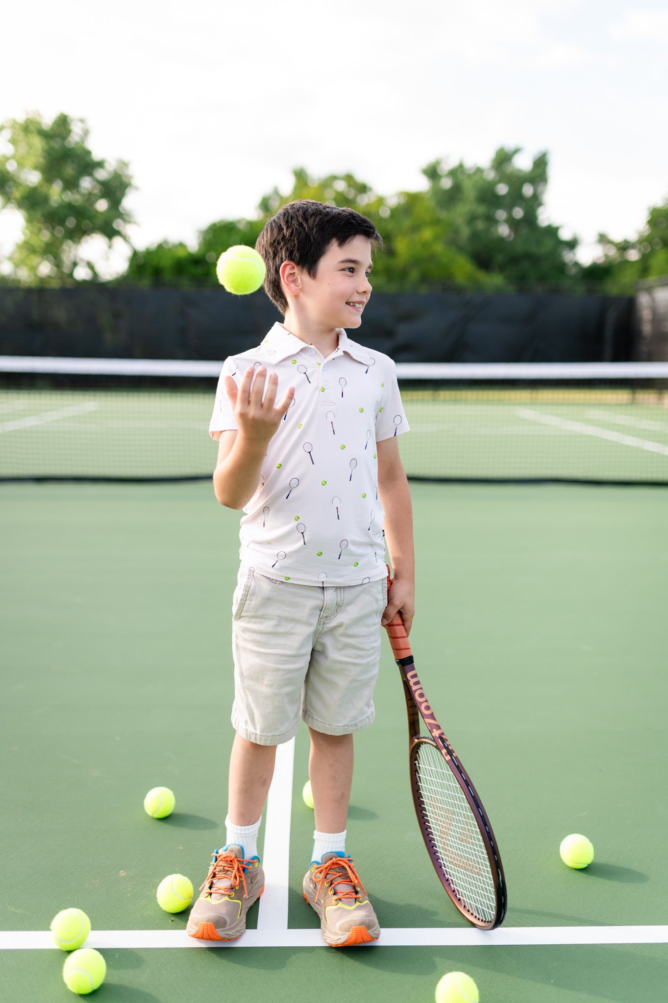 Child on a tennis court with a racket and ball wearing a polo short sleeve top with tennis pattern, surrounded by tennis balls.