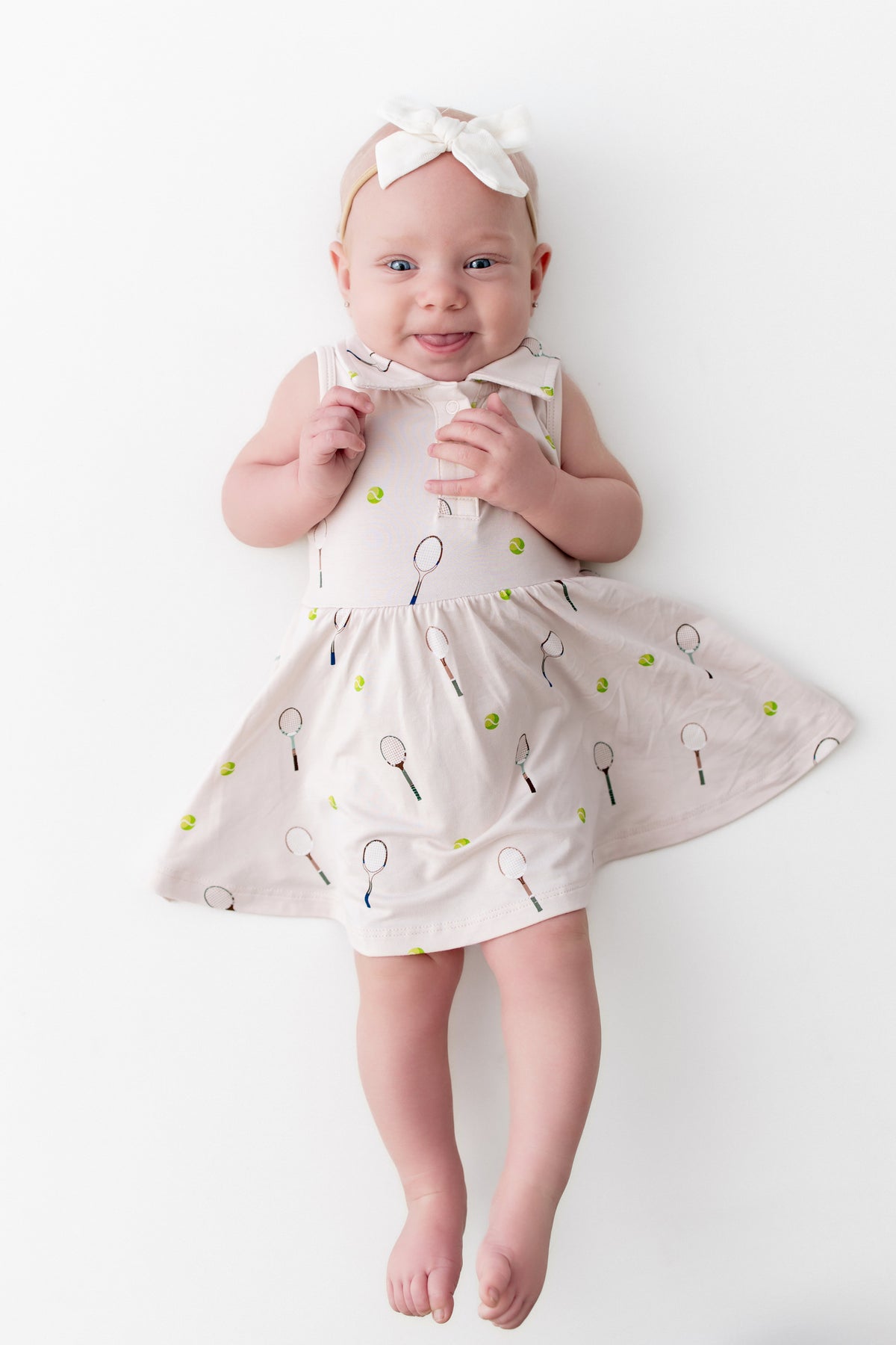 Baby wearing a white dress with tennis racket and green tennis ball pattern on a white background